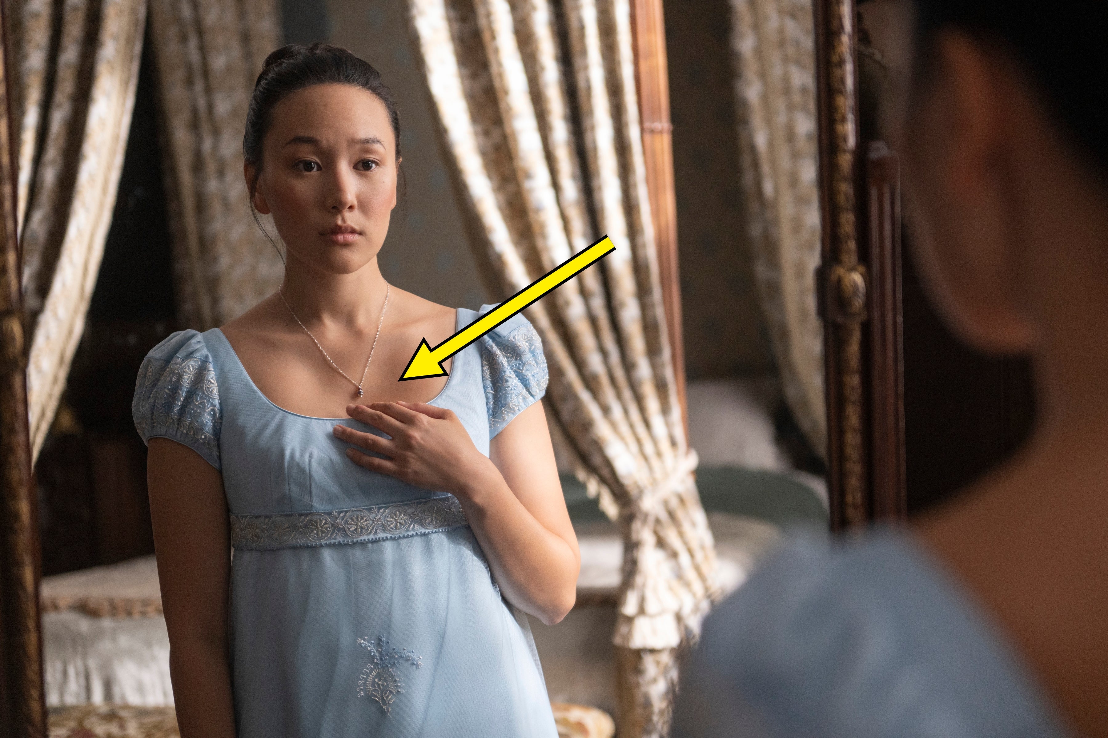 Person wearing Regency-style dress looks thoughtfully at their reflection in a mirror, standing in a room with draped curtains