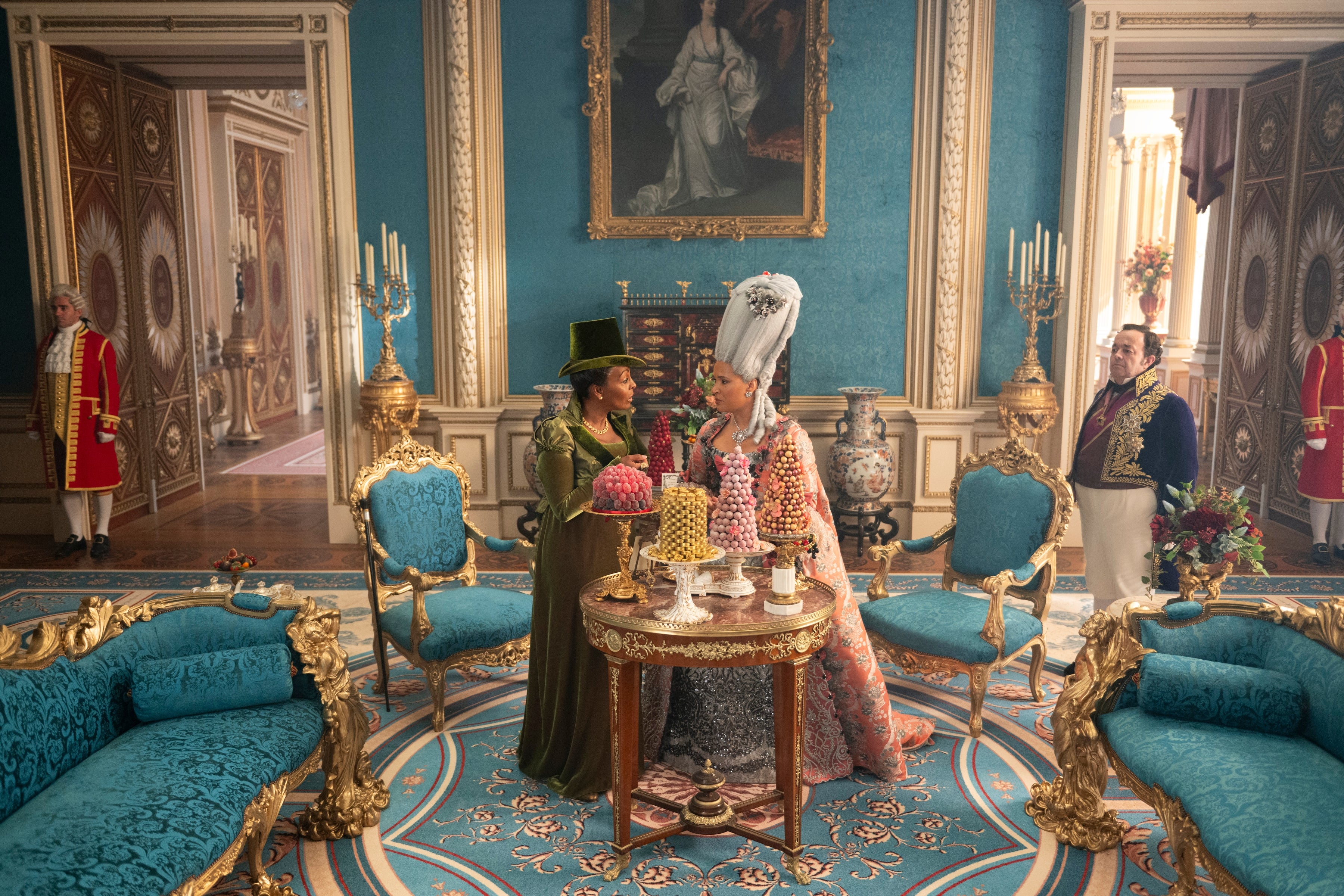 Two people in elaborate period costumes converse over a table with desserts in an opulent room. Background features ornate decor and attendants