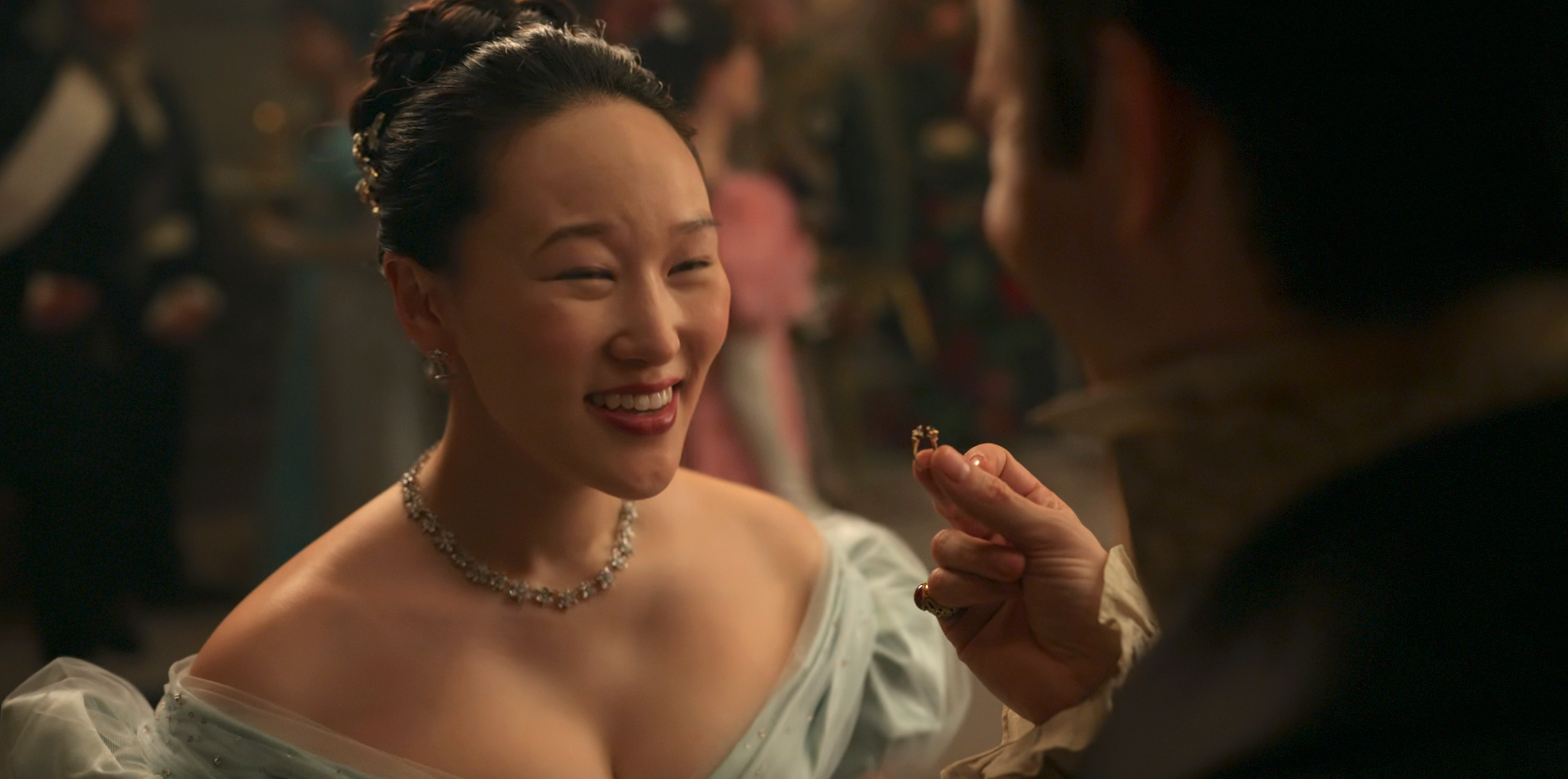 A woman in an elegant off-shoulder gown smiles at a man holding something small in a social gathering setting