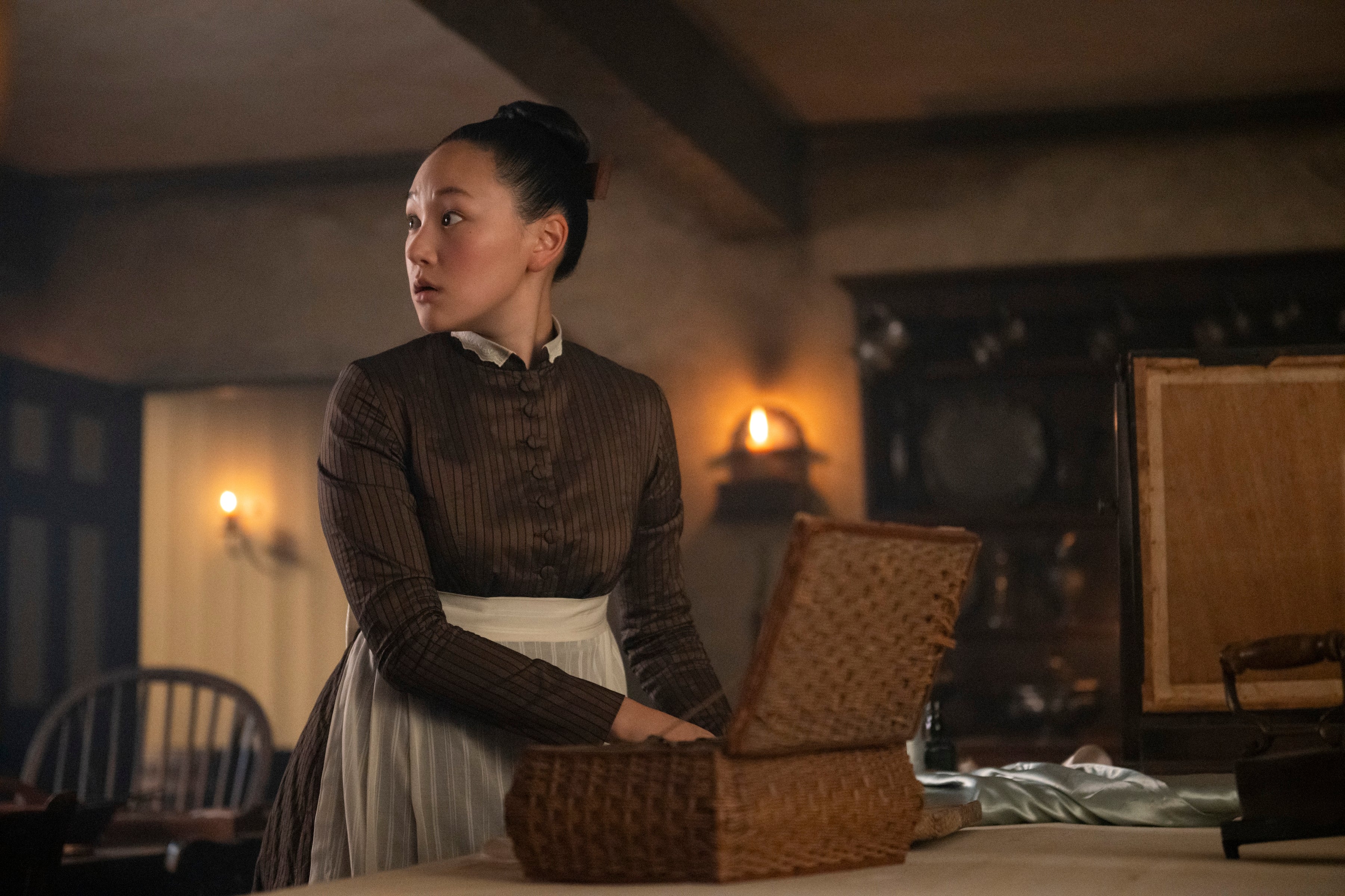 A person in period clothing stands by a table with a woven basket in a dimly lit room, looking off to the side