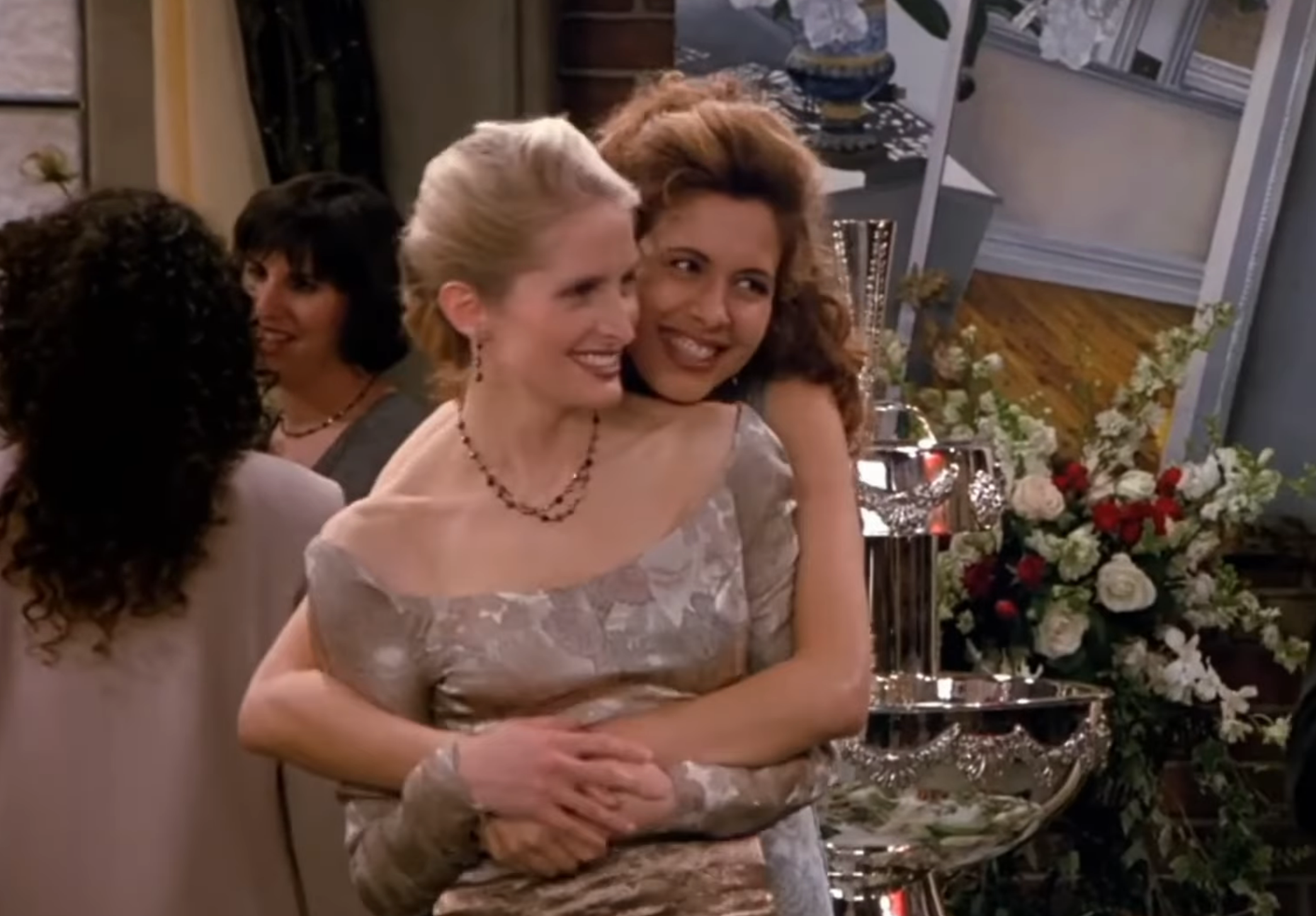 Two radical   successful  elegant attire are embracing astatine  a ceremonial  event. One is smiling broadly, portion    the different   is looking content. A decorative inheritance  is visible