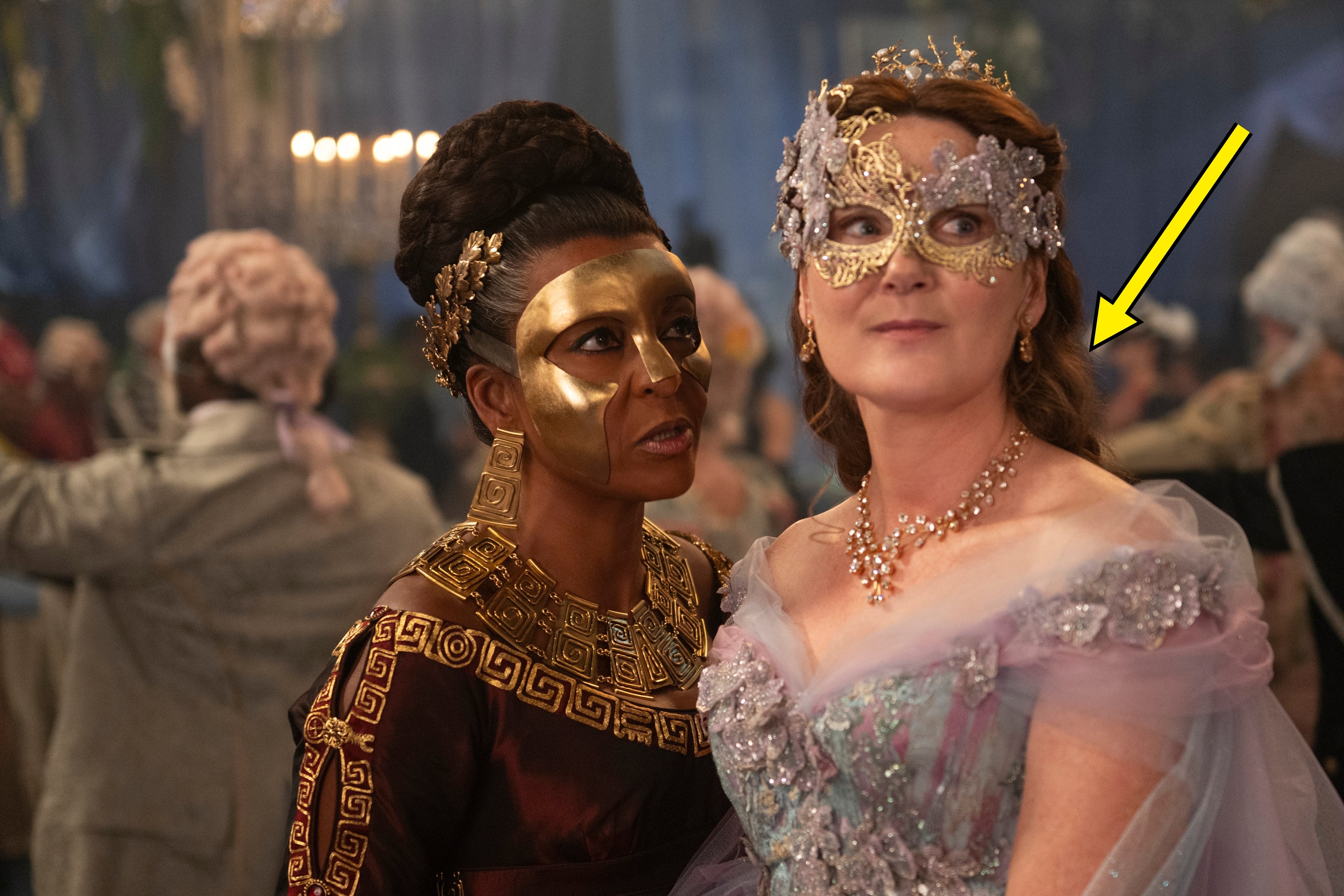 Two people wearing detailed costumes and ornate masks at a masquerade ball, engaging in conversation amidst a blurred festive background