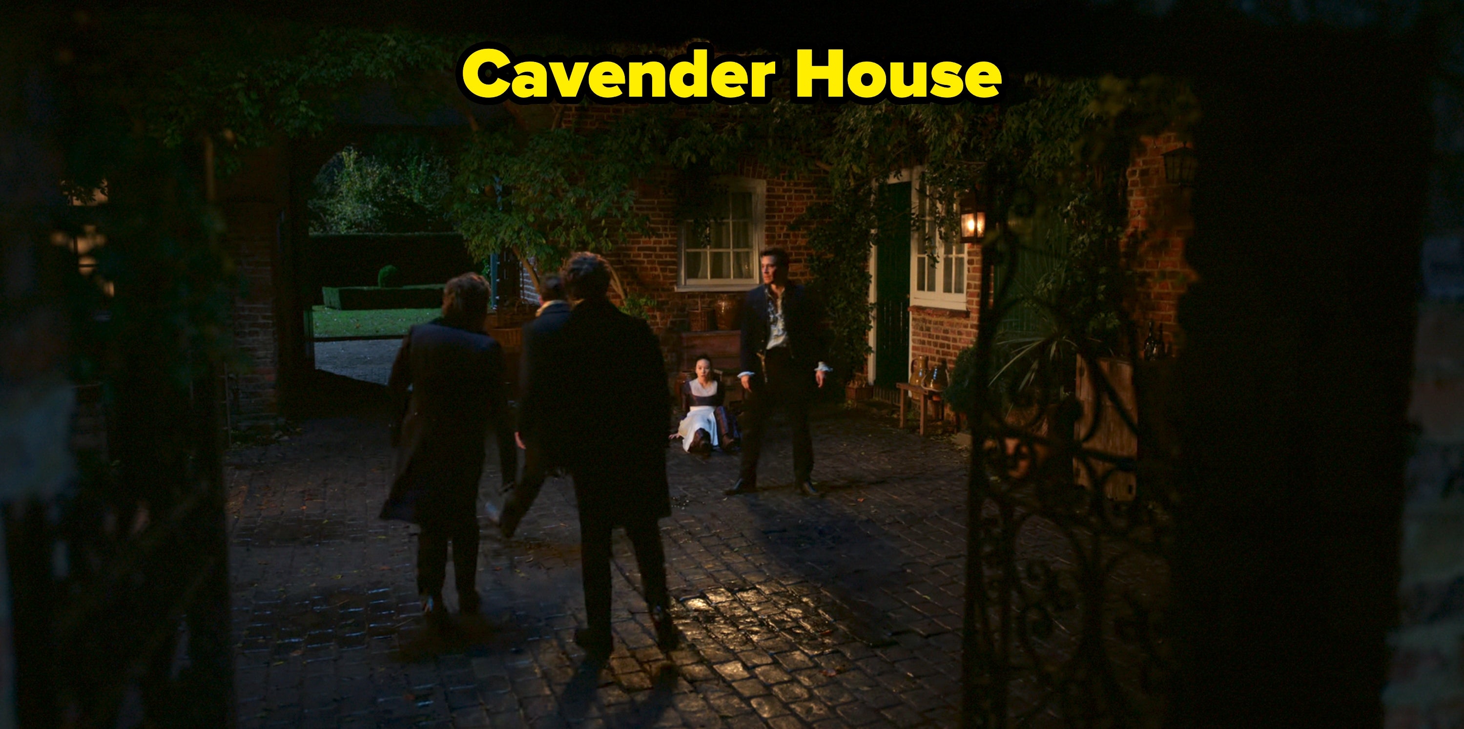 Characters in formal attire confront a person sitting on the cobblestone ground in a dimly lit courtyard scene