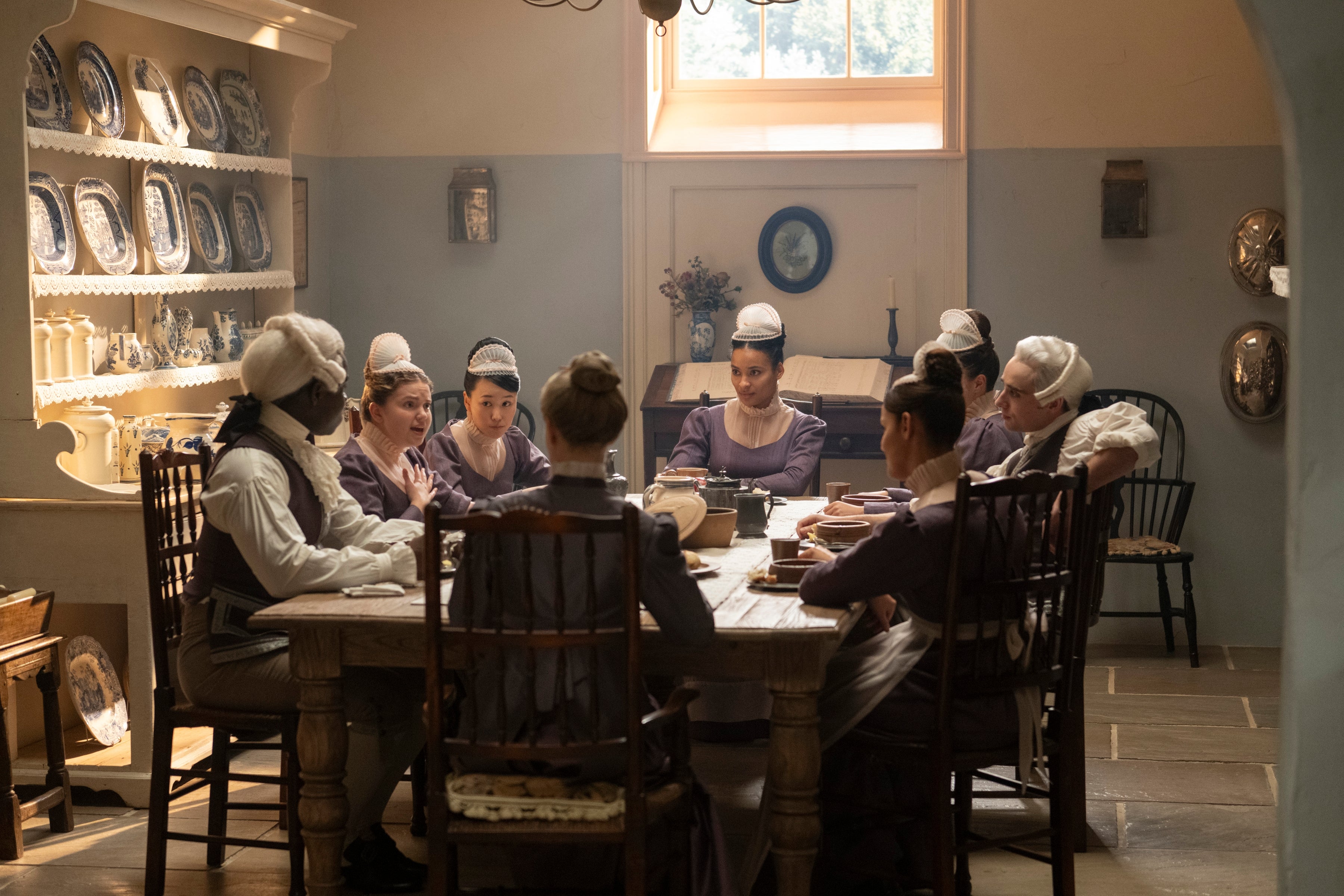 People dressed in historic attire sit around a table in a period drama setting