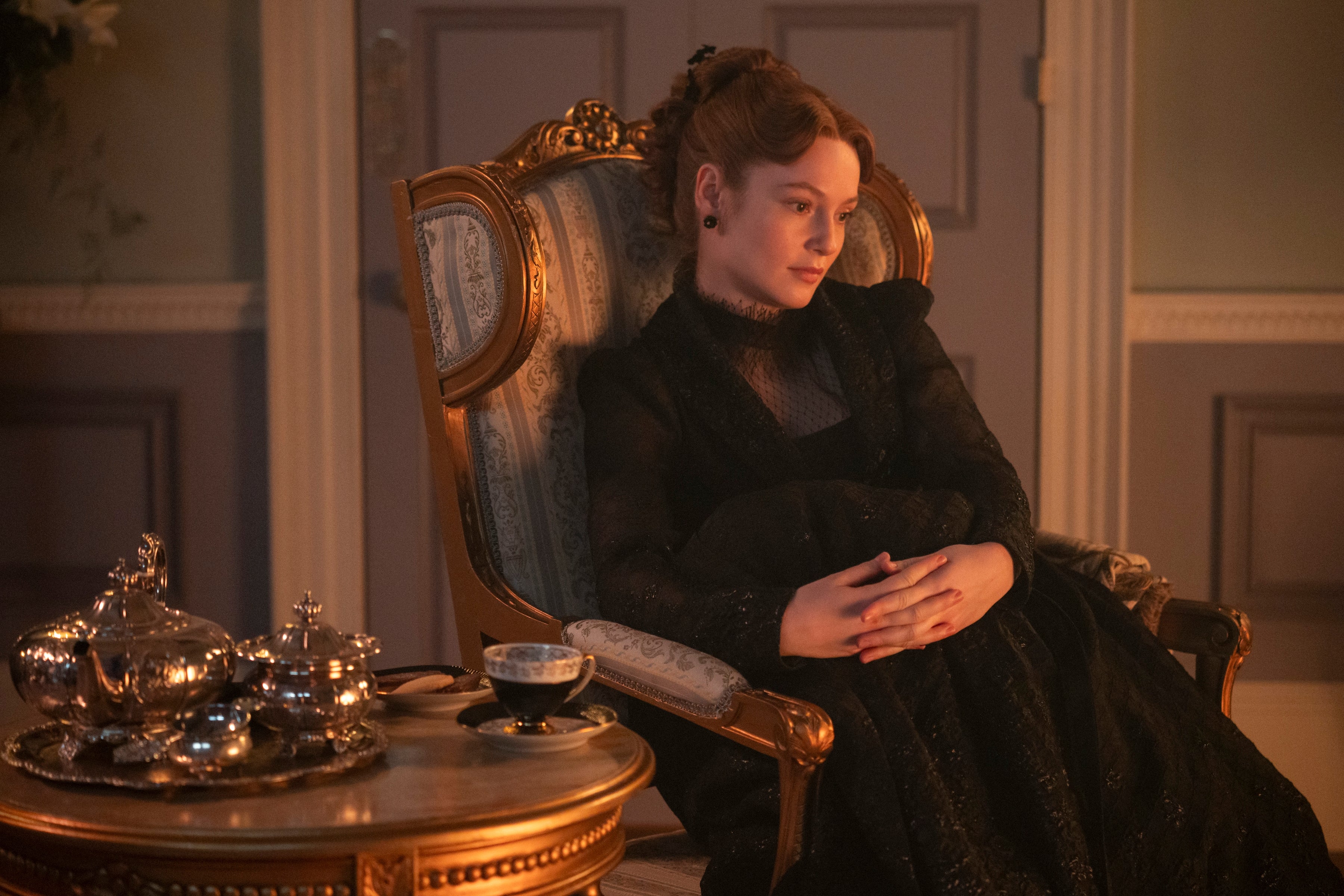 Person in period costume, with intricate lace details, sits thoughtfully in an ornate chair next to a silver tea set, evoking a historical setting