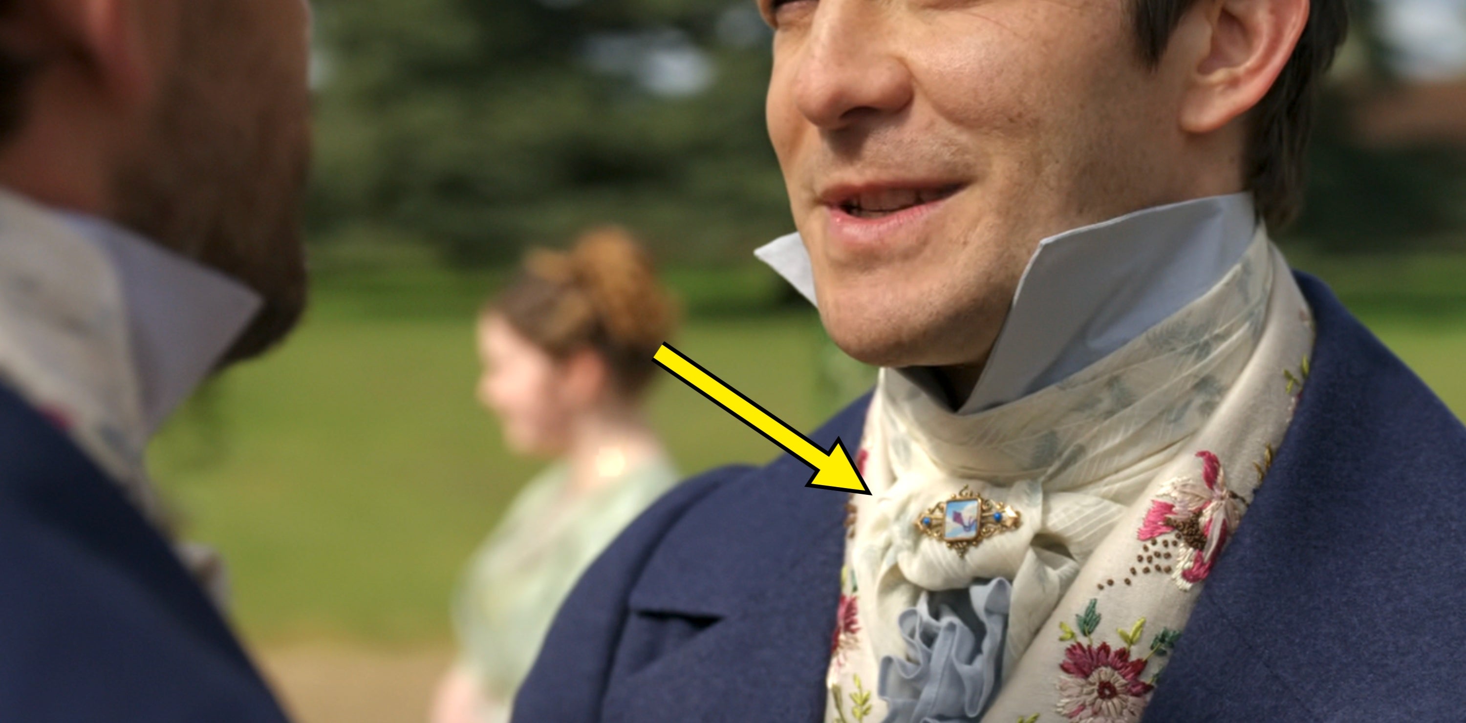 Two men in historical costume with patterned cravats converse outdoors, reminiscent of period dramas