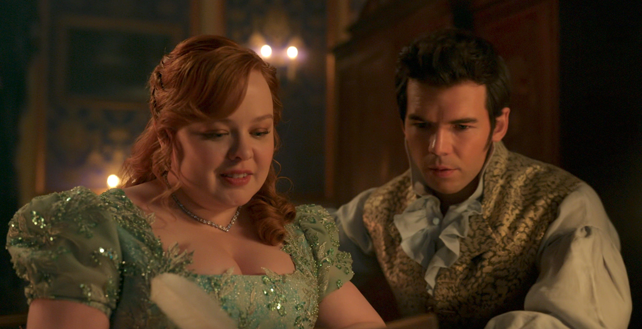 Two actors in a period drama setting, one in an ornate dress and the other in a vintage vest, sit together looking intently at a document