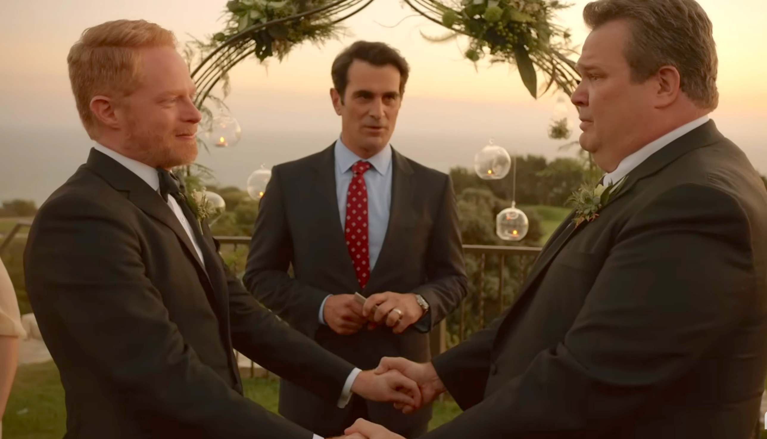 Two men successful  suits clasp  hands astatine  a wedding ceremony, with different  antheral   successful  a suit   officiating the nuptials outdoors