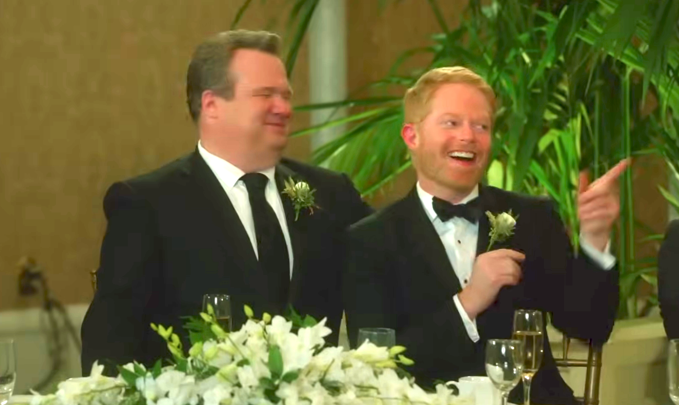 Two radical   successful  achromatic  suits are sitting astatine  a array  with achromatic  flowers, smiling and engaged successful  speech  astatine  a ceremonial  event