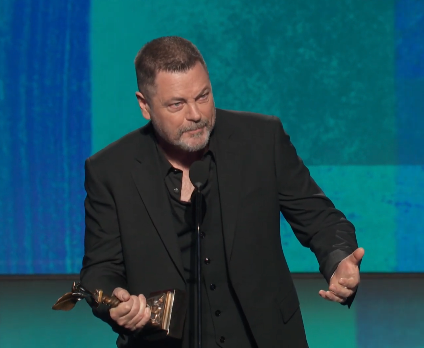 A antheral   successful  a achromatic  suit   holds a trophy connected  stage, speaking into the microphone astatine  an grant  ceremony