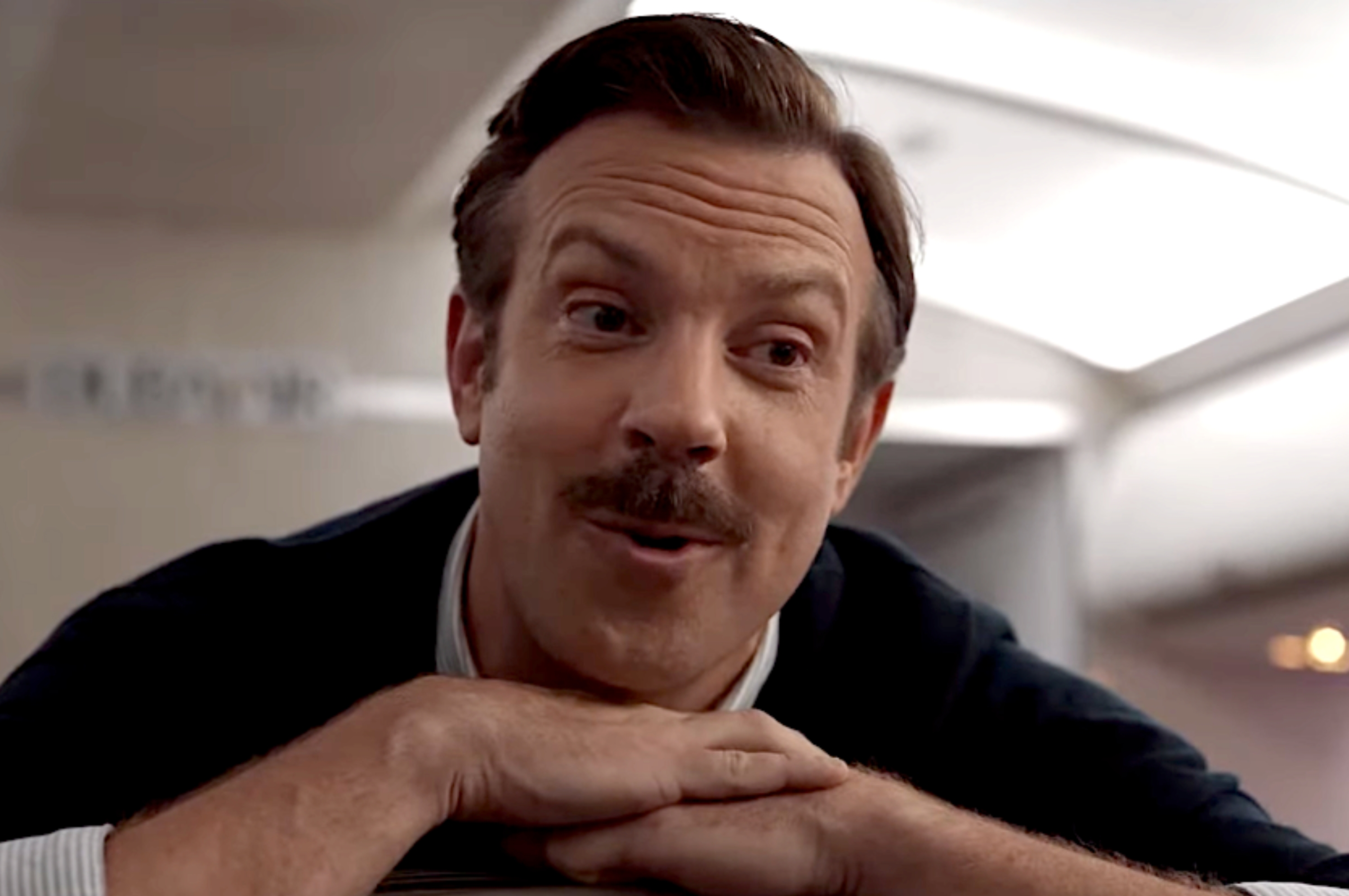 Man with a mustache, wearing a striped shirt and sweater, leans on airplane seat with an amused expression in an aircraft cabin
