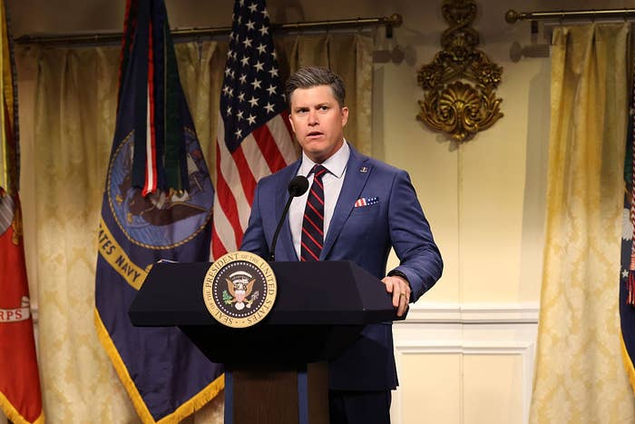 Person in a suit speaks at a podium with a presidential seal, flanked by flags. Background features decorative elements