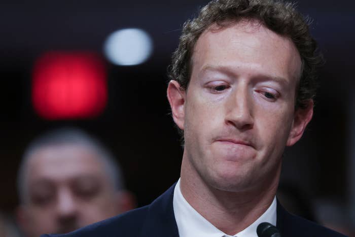 Mark Zuckerberg in a suit appears contemplative or tense while surrounded by onlookers, likely at a formal or important event