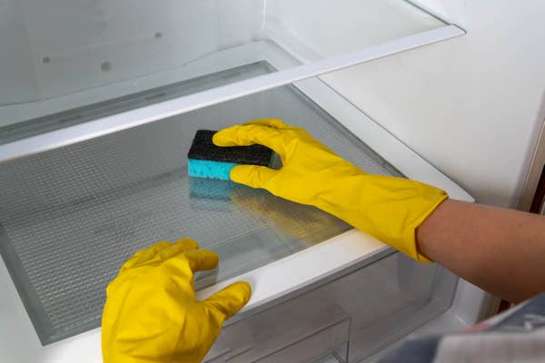 Person wearing yellow gloves cleaning a fridge shelf with a sponge
