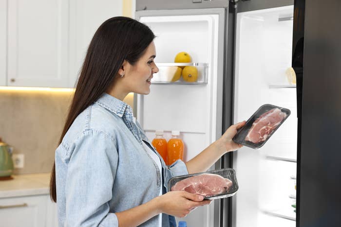 Woman in kitchen holding packaged meat, looking into a refrigerator