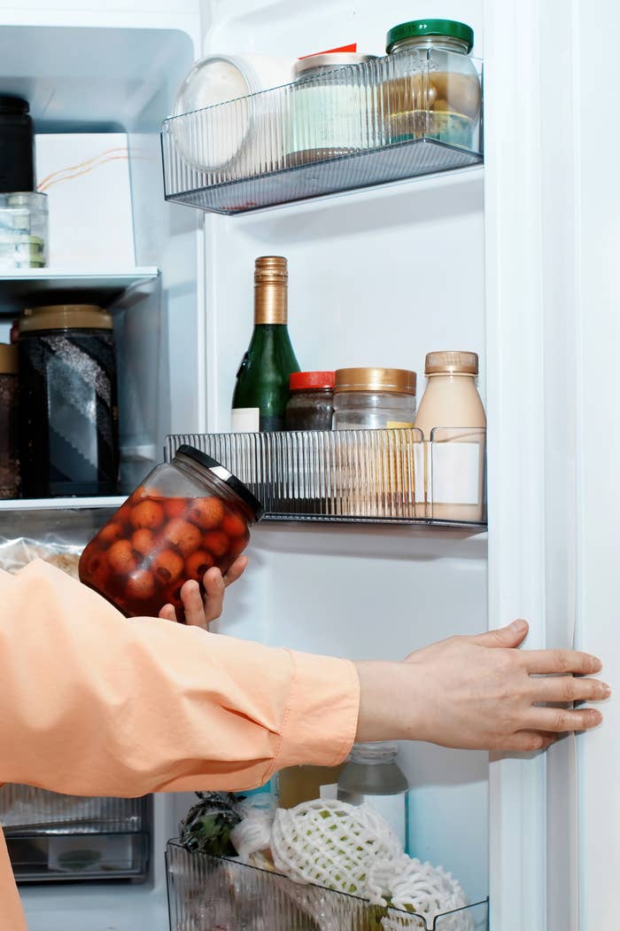 Person in an orange shirt placing a jar of preserved food into a refrigerator door shelf filled with various bottles and jars