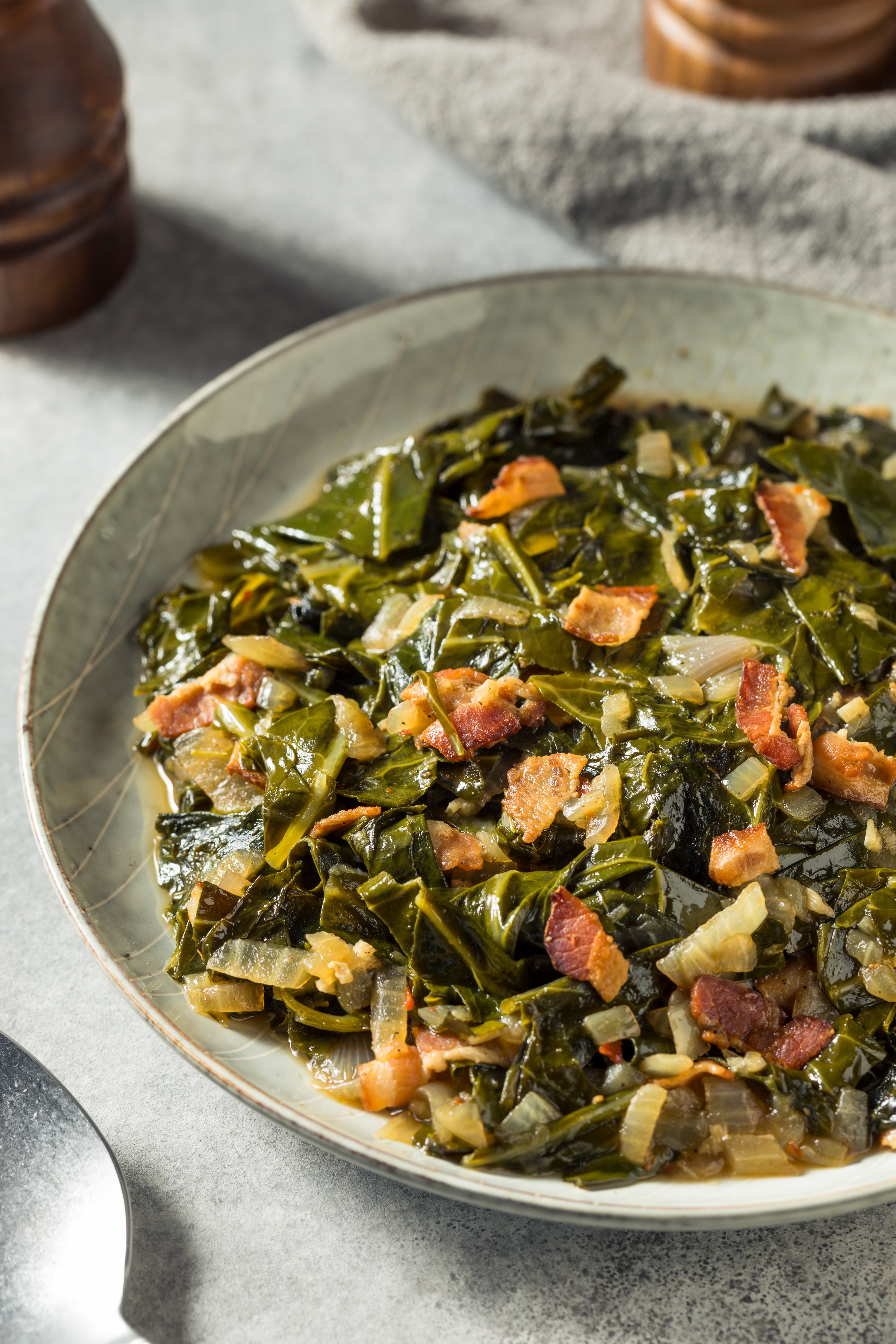 A vessel  of collard greens mixed with pieces of bacon and onions connected  a table, adjacent to a spoon and capsicum  mill
