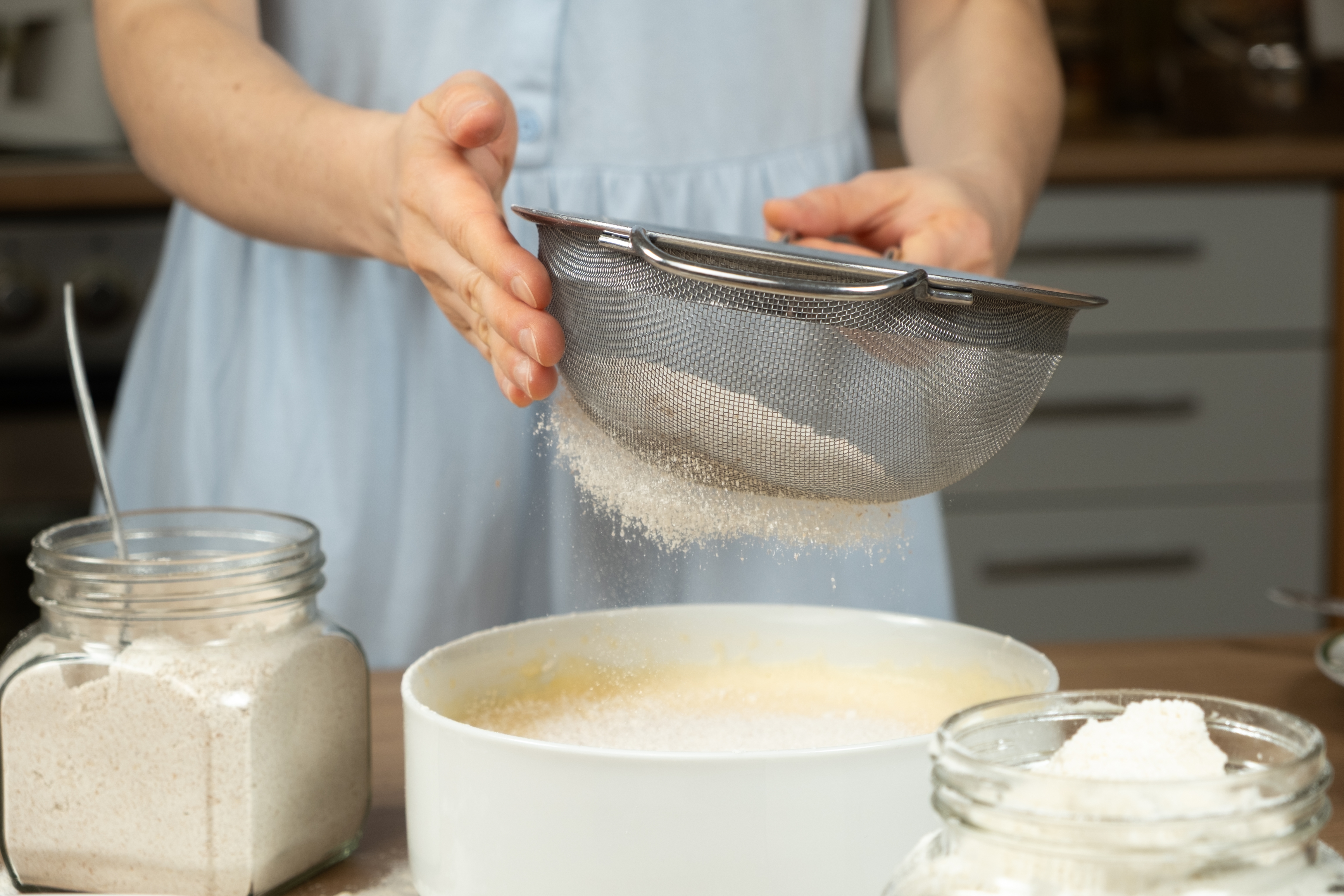 Person sifting flour into a vessel with jars of ingredients connected a room counter