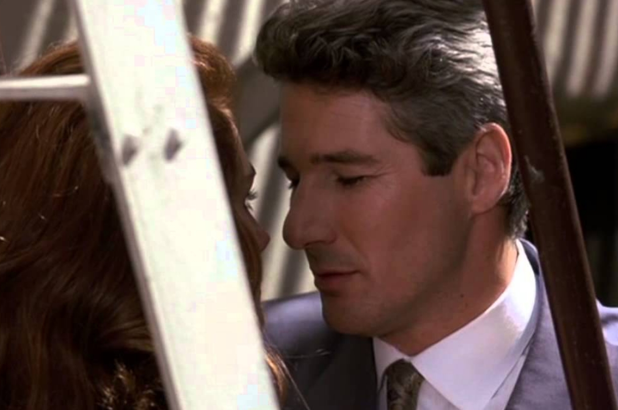 A couple shares an intimate moment, standing closely together, partially obscured by a ladder. The man wears a suit