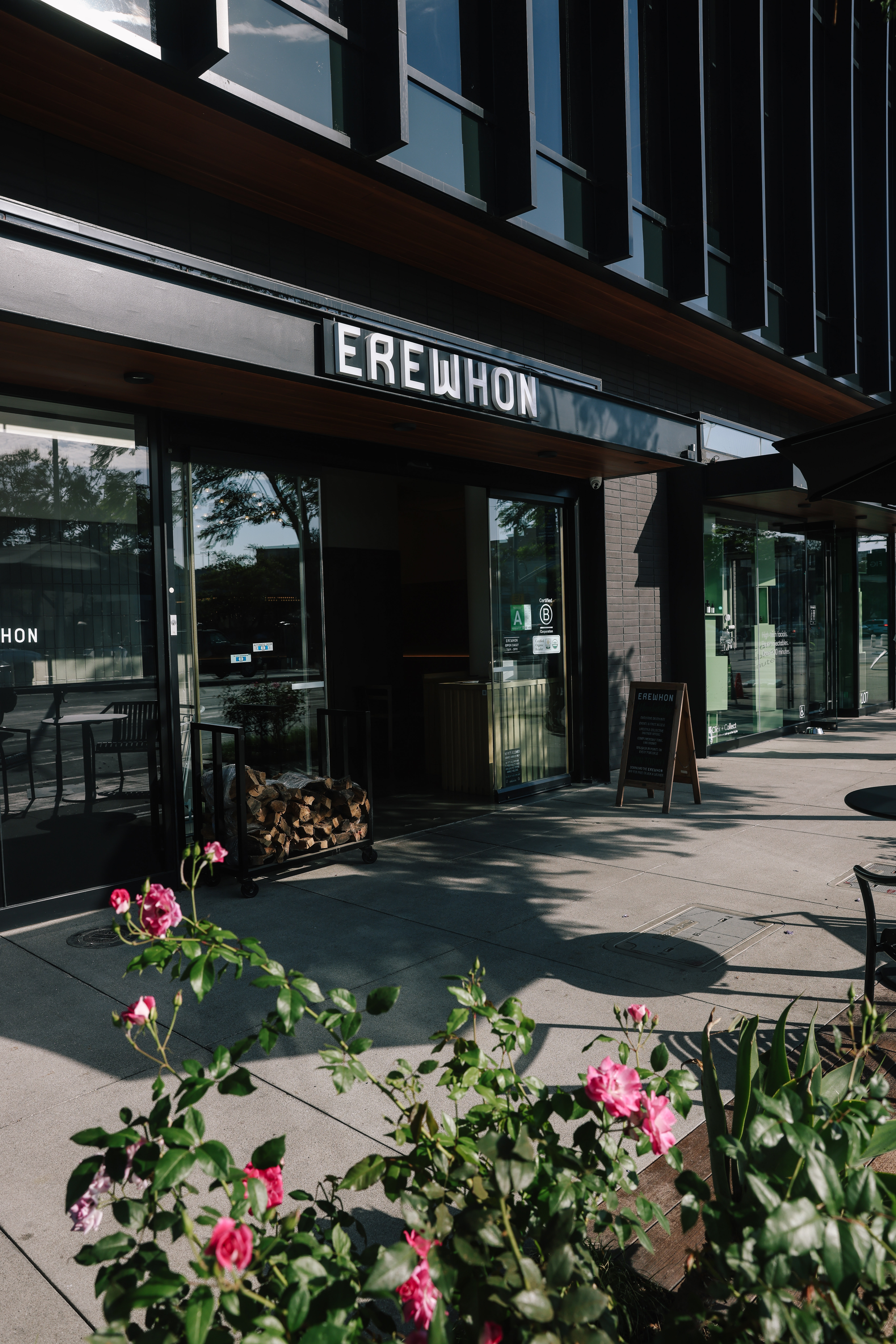 Storefront with "Erewhon" signage supra  the entrance, outdoor seating visible. Flowers successful  the foreground, shadows formed  connected  the pavement