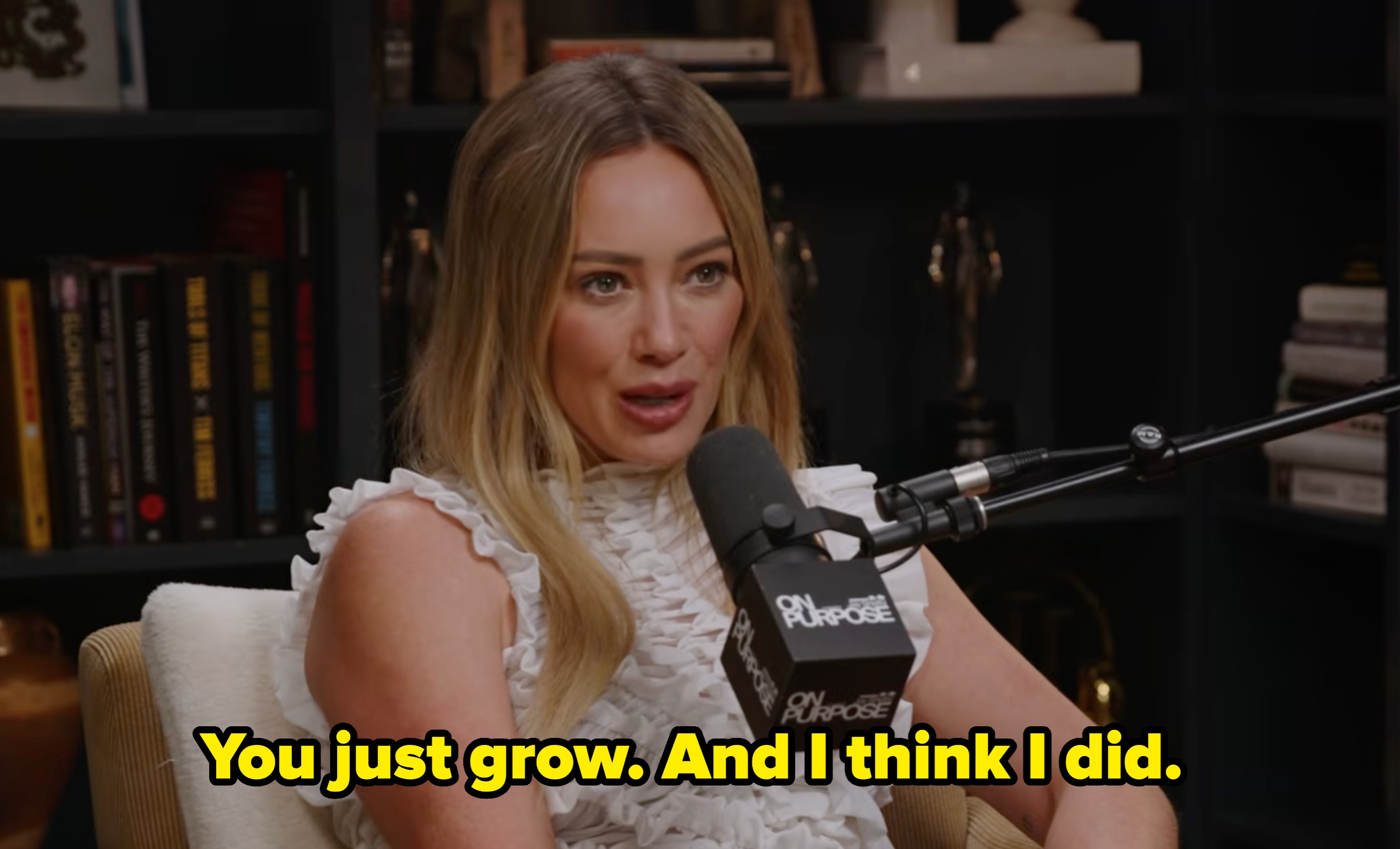 Person speaking into a microphone during a podcast. They wear a ruffled sleeveless top. Books and awards are visible in the background