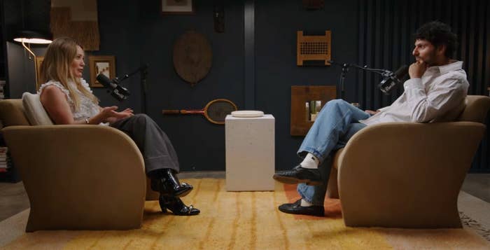 Two people sit in armchairs having a podcast-style conversation. They have microphones in front of them