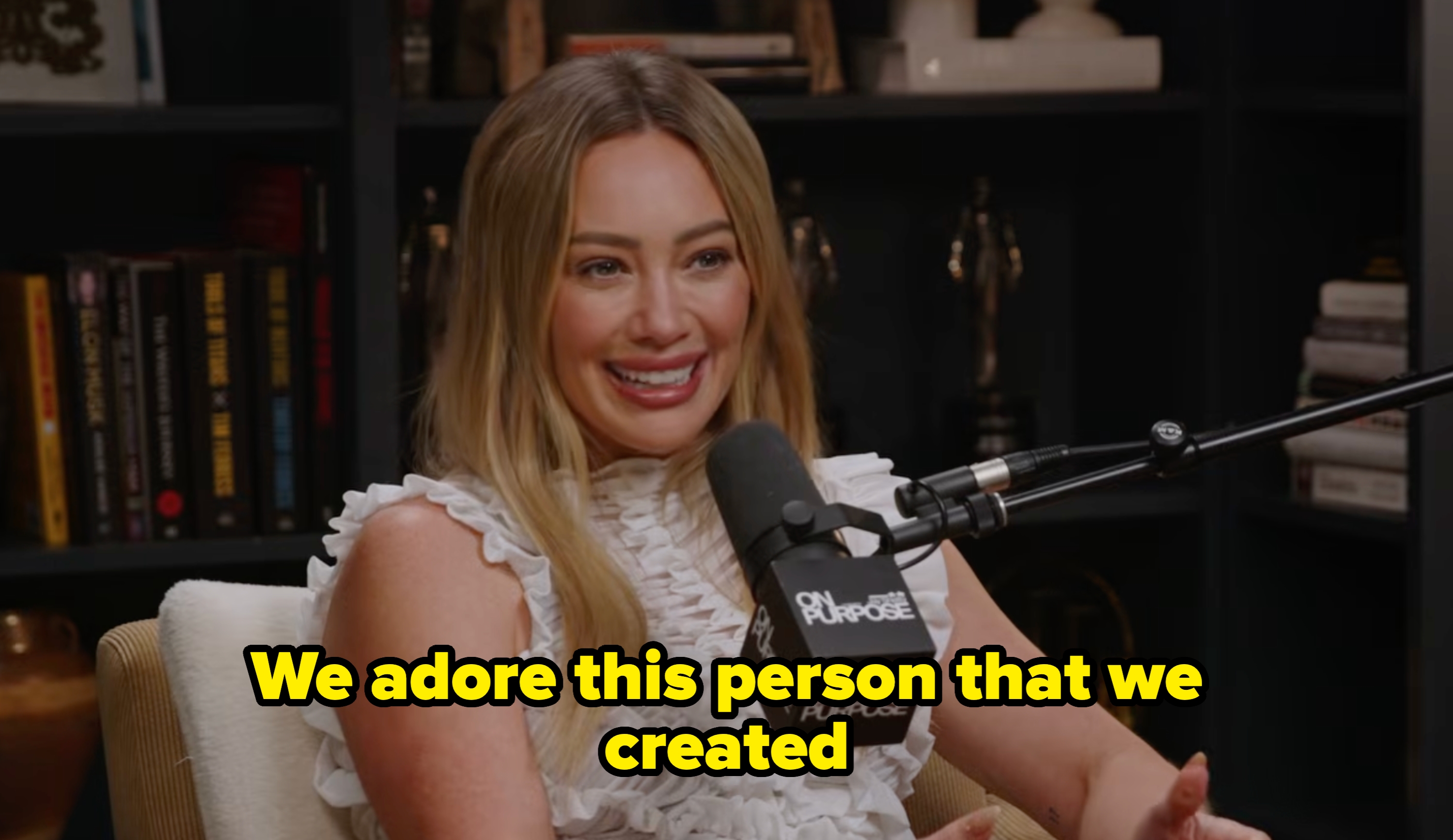 Person smiling during a podcast interview, wearing a ruffled top, seated with a microphone in front of them