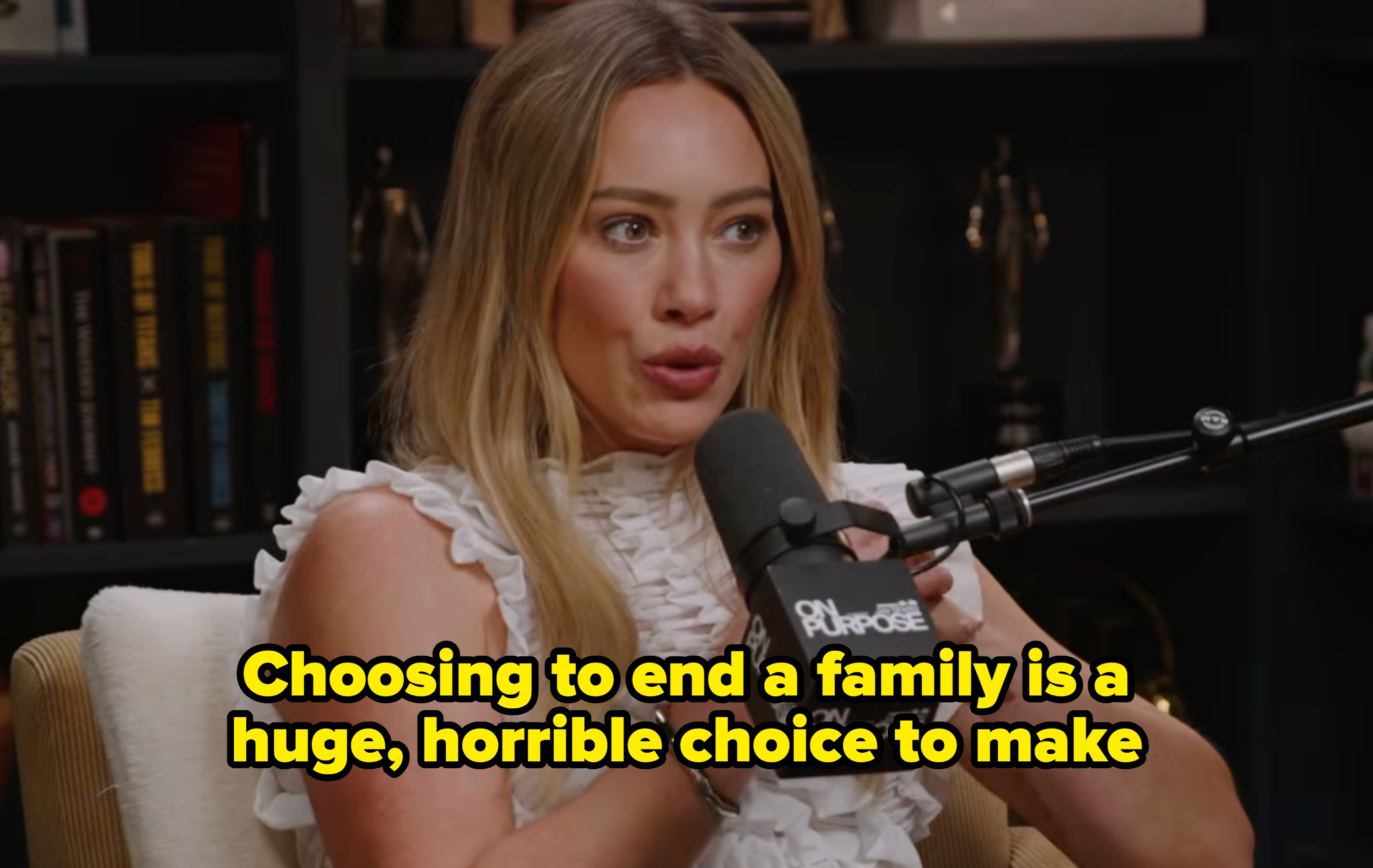 Person in ruffled top speaking into a microphone on a podcast set, with books and awards in the background