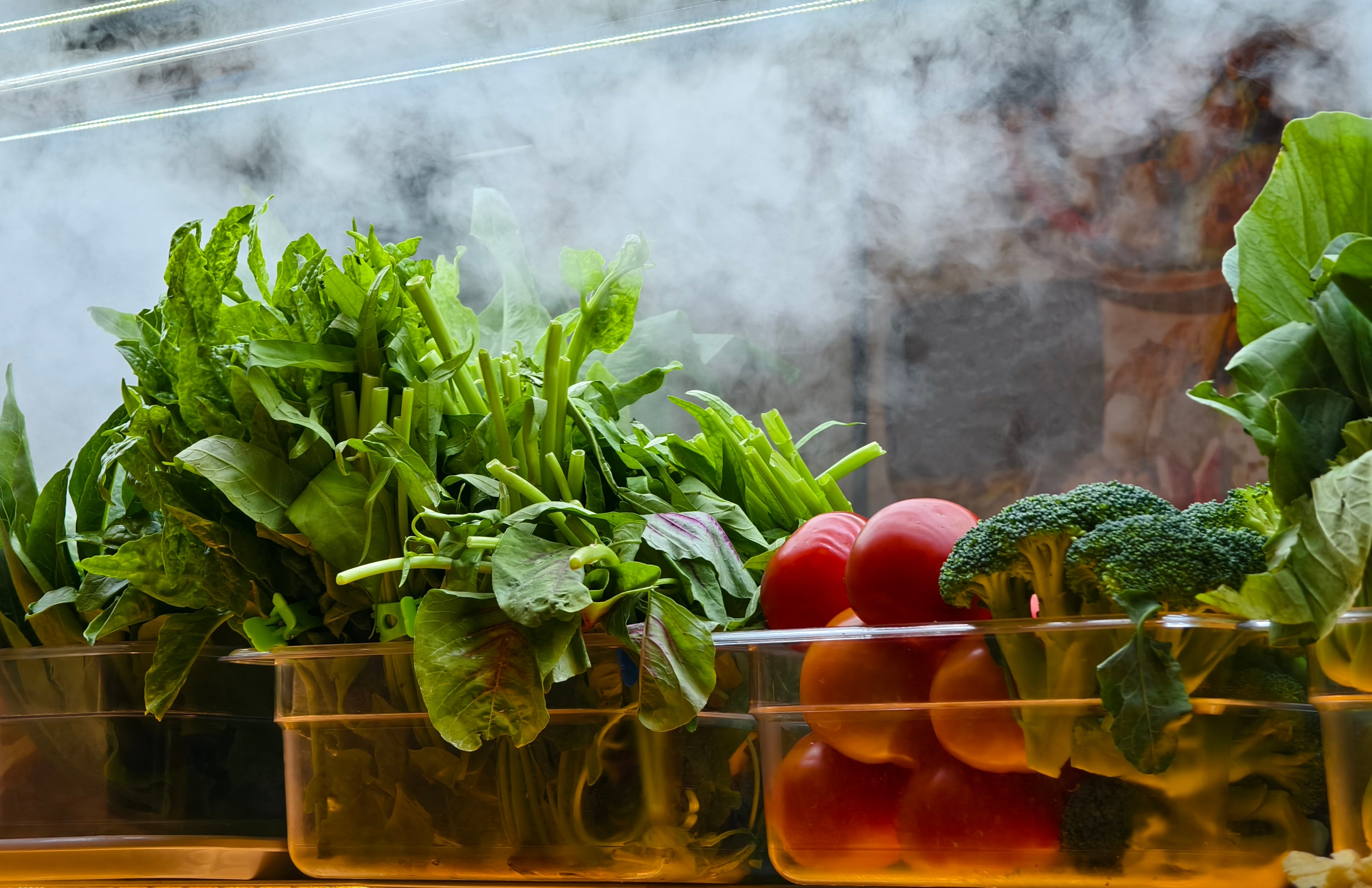 Bins of fresh greens, tomatoes, and broccoli on display with steam in the background