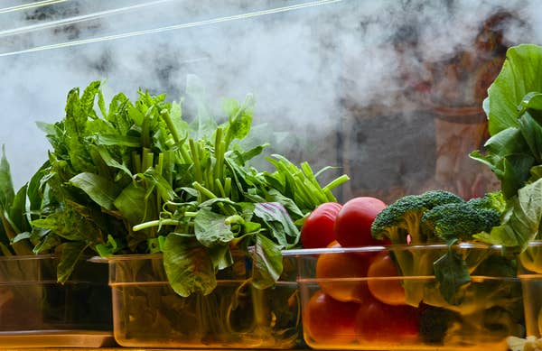 Bins of fresh greens, tomatoes, and broccoli on display with steam in the background
