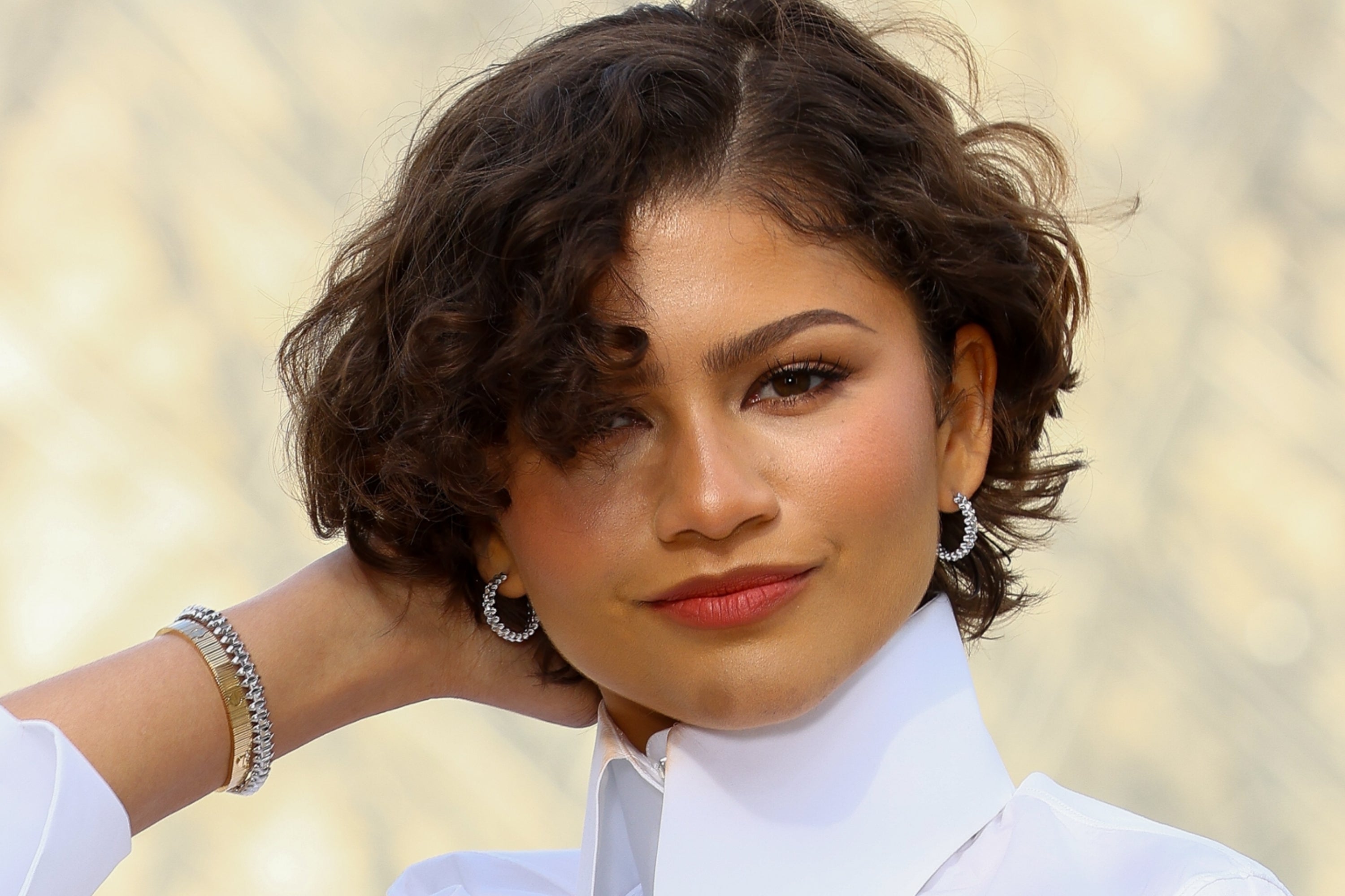 Person in a high-collared white shirt poses confidently with a hand behind their head, wearing hoop earrings and a bracelet