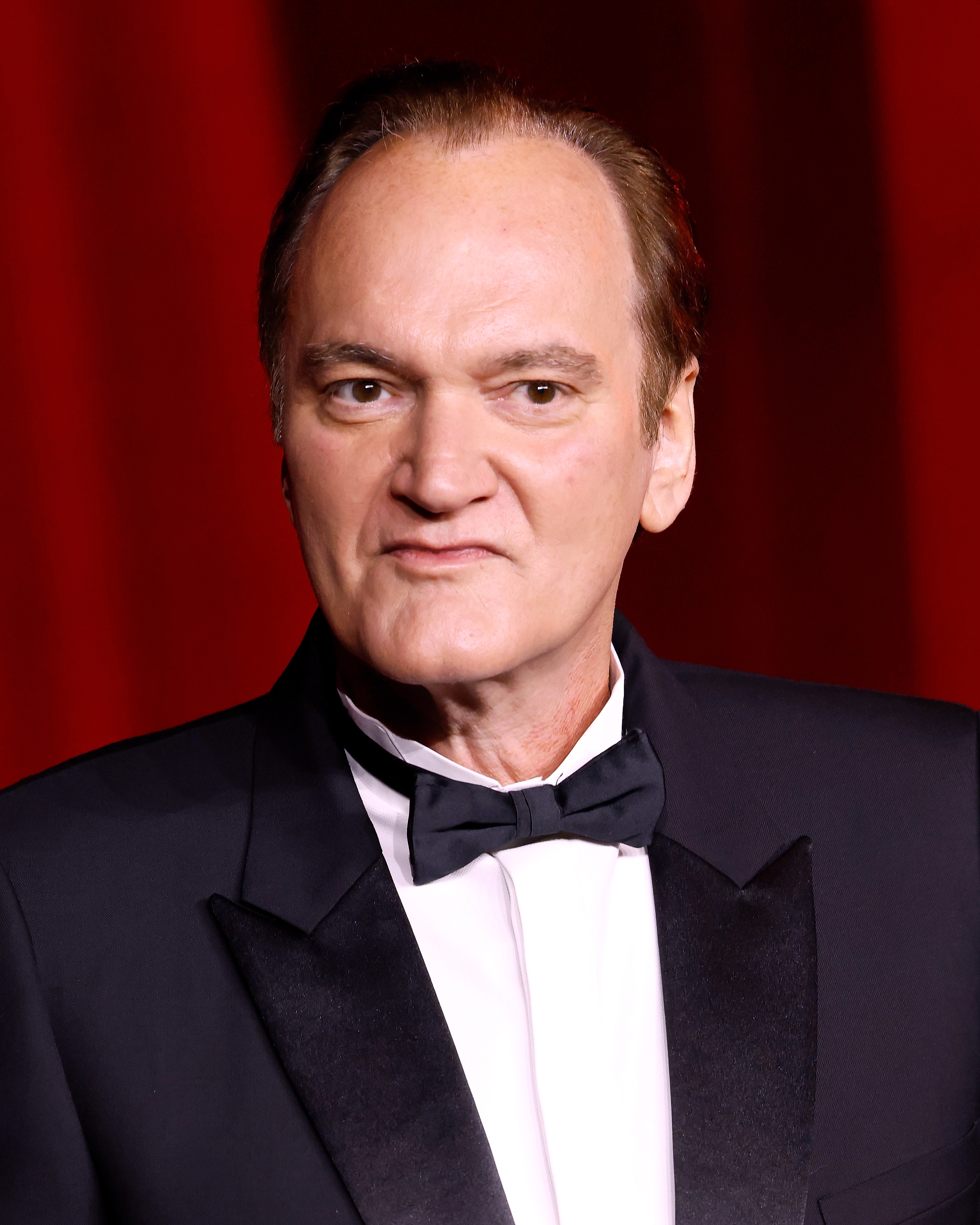 Man successful  a classical  tuxedo with a bow necktie  stands against a reddish  backdrop astatine  a ceremonial  event