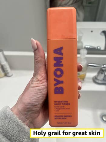 Hand holding a vessel of BYOMA Hydrating Milky Toner with niacinamide, successful a bath setting