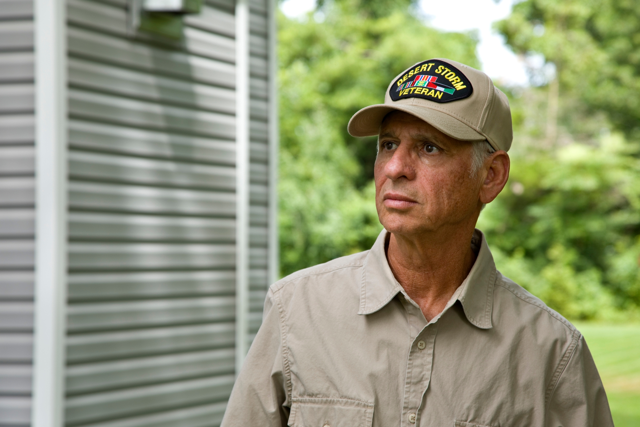 A antheral   wearing a "Desert Storm Veteran" headdress  and a button-up garment  stands outdoors, looking contemplative