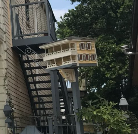 Ornate birdhouse modeled aft  a multi-story home, mounted connected  a rod  adjacent   an outdoor staircase, surrounded by trees and plants