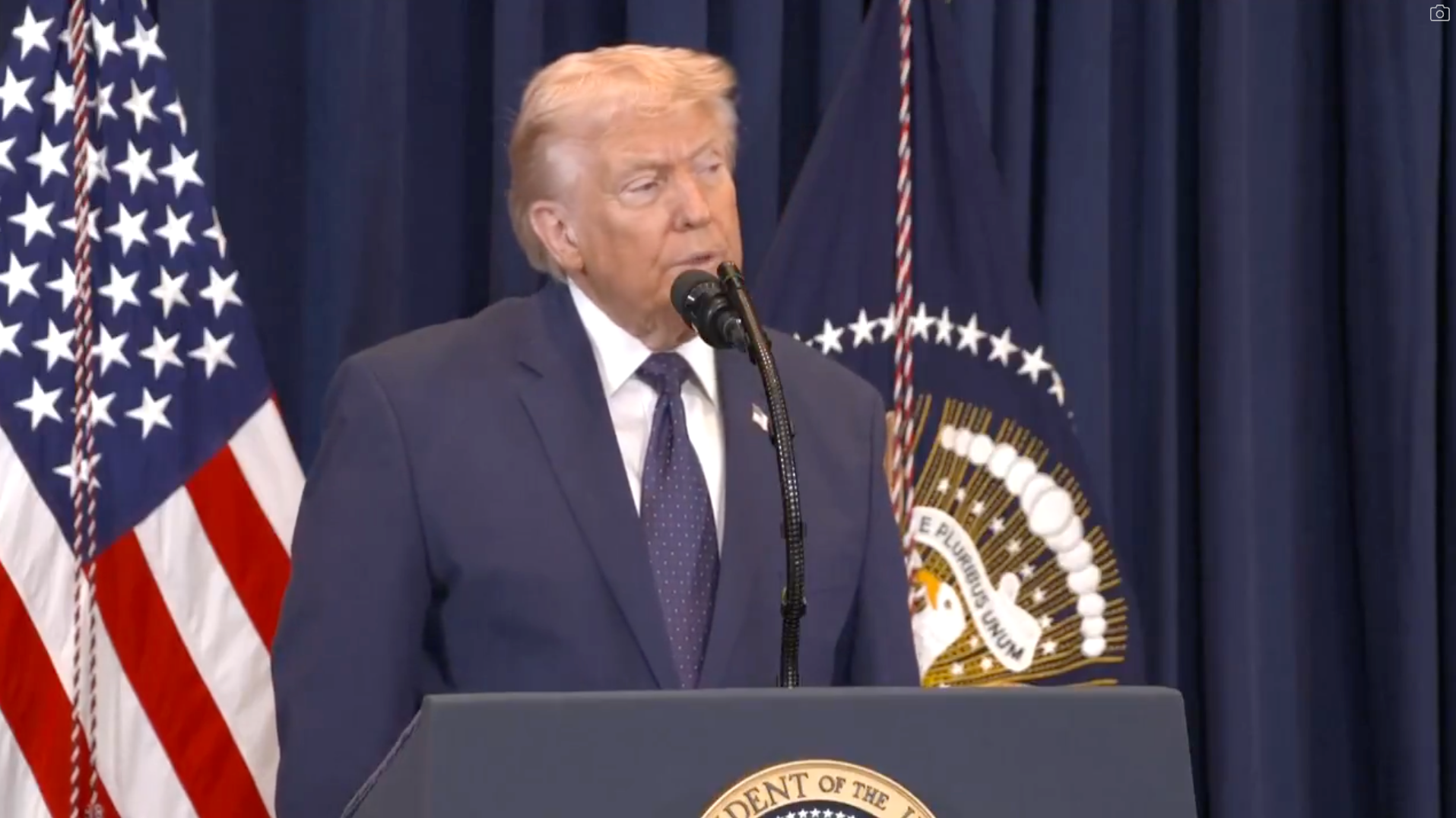 En mann i dress taler ved et podium med amerikanske flagg og et presidentstempel i bakgrunnen, ved en formell begivenhet