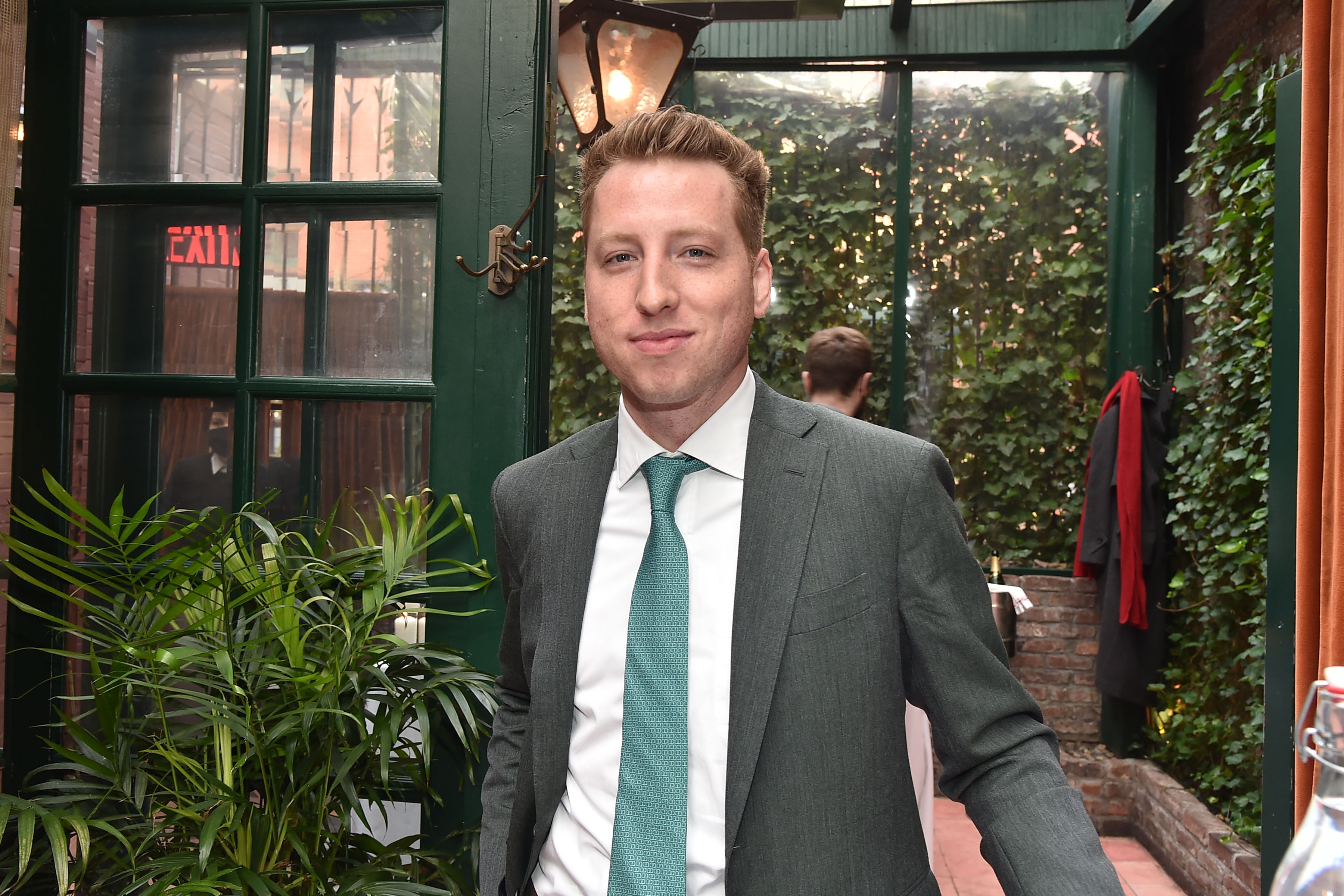 Person i dress og slips står i et elegant dekorert innendørs hageområde, med grøntområder og en lysarmatur i lanternestil