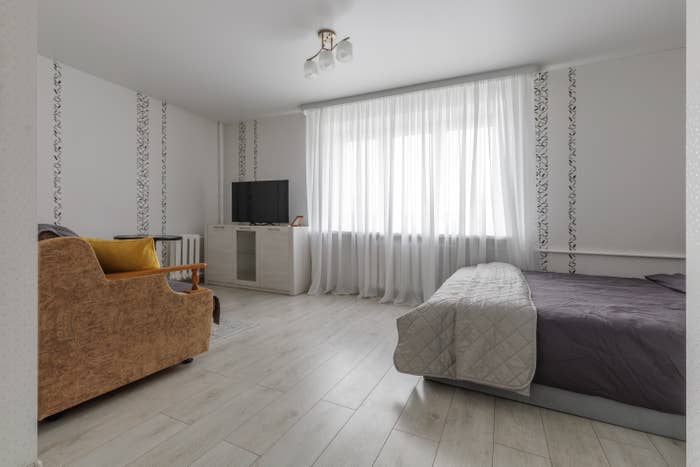 Minimalist chamber  with a bed, TV connected  a stand, and a brownish  sofa. Sheer curtains screen  a ample  window, letting successful  earthy  light