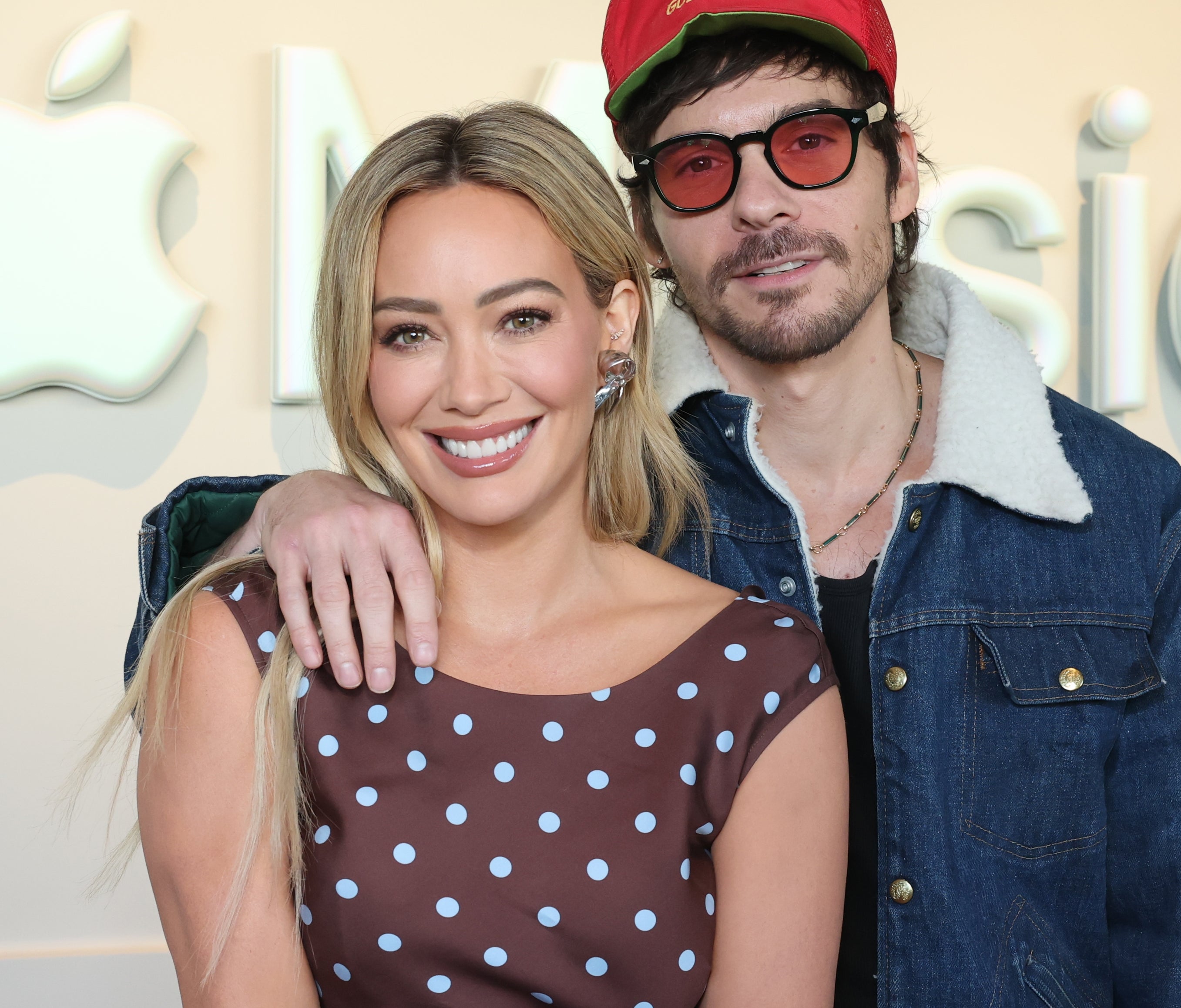 Two people smiling at an Apple Music event. One wears a polka dot dress, the other a jacket with a fur collar and red hat