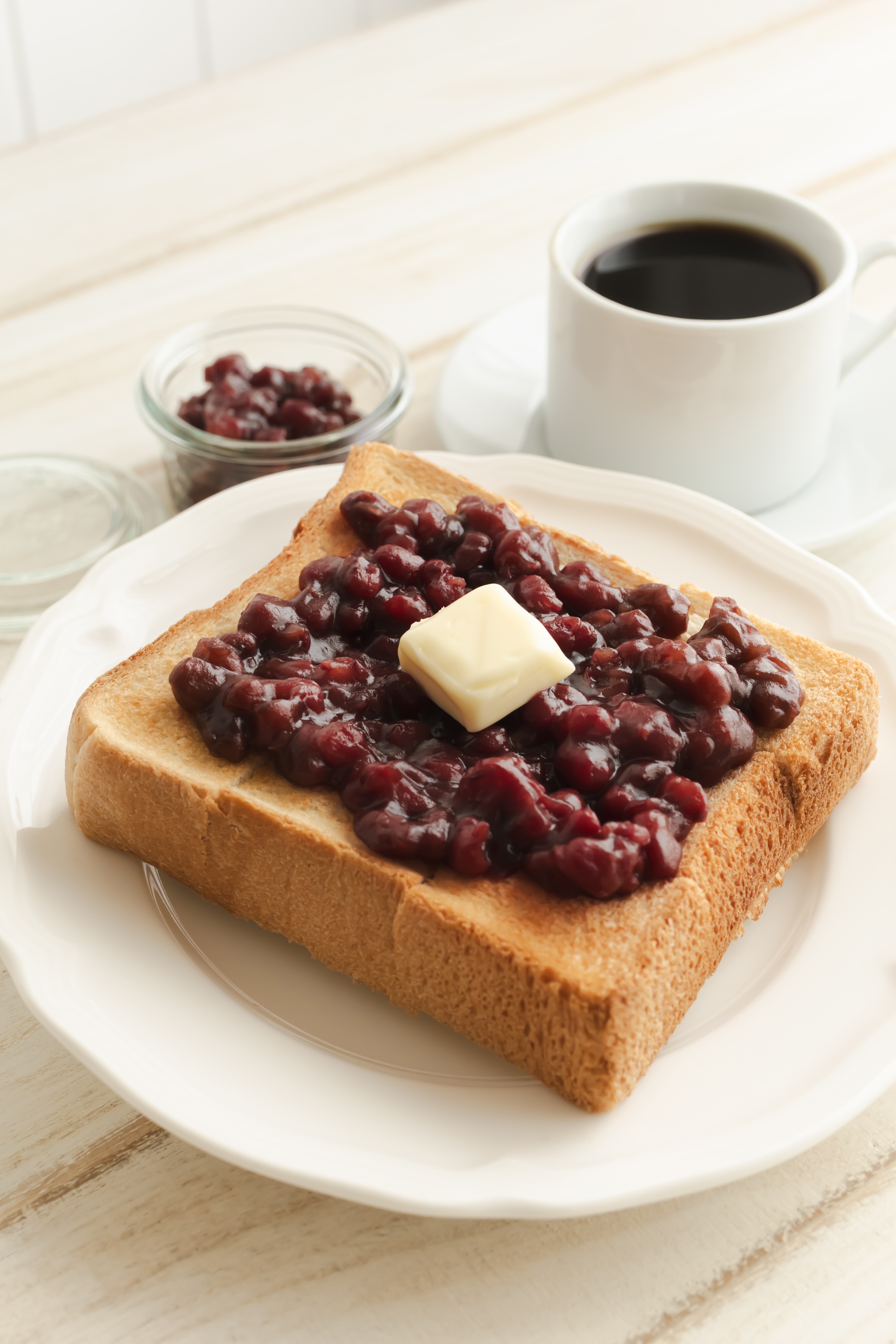 厚切りトーストにたっぷりのあんことバターがのっている。後ろにはコーヒーカップと小瓶に入ったあんこ。