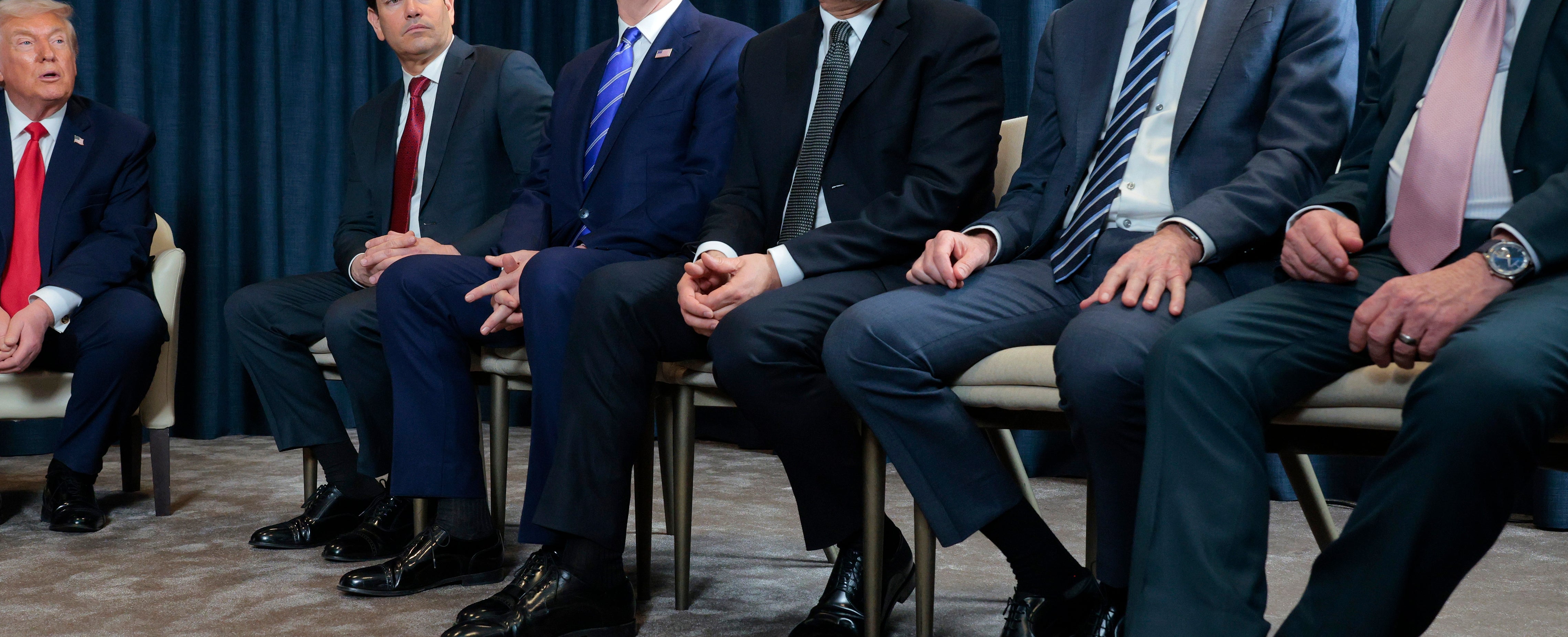 A group of men in suits seated in a conference room, with a NATO flag in the background, but a close up of their feet and nearly all of them are wearing identical shoes