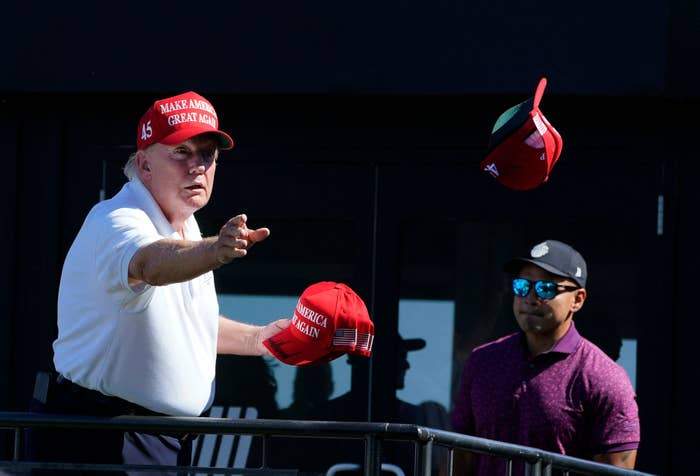 Donald Trump in a polo shirt tosses a "Make America Great Again" cap while another person stands nearby wearing sunglasses and a golf shirt