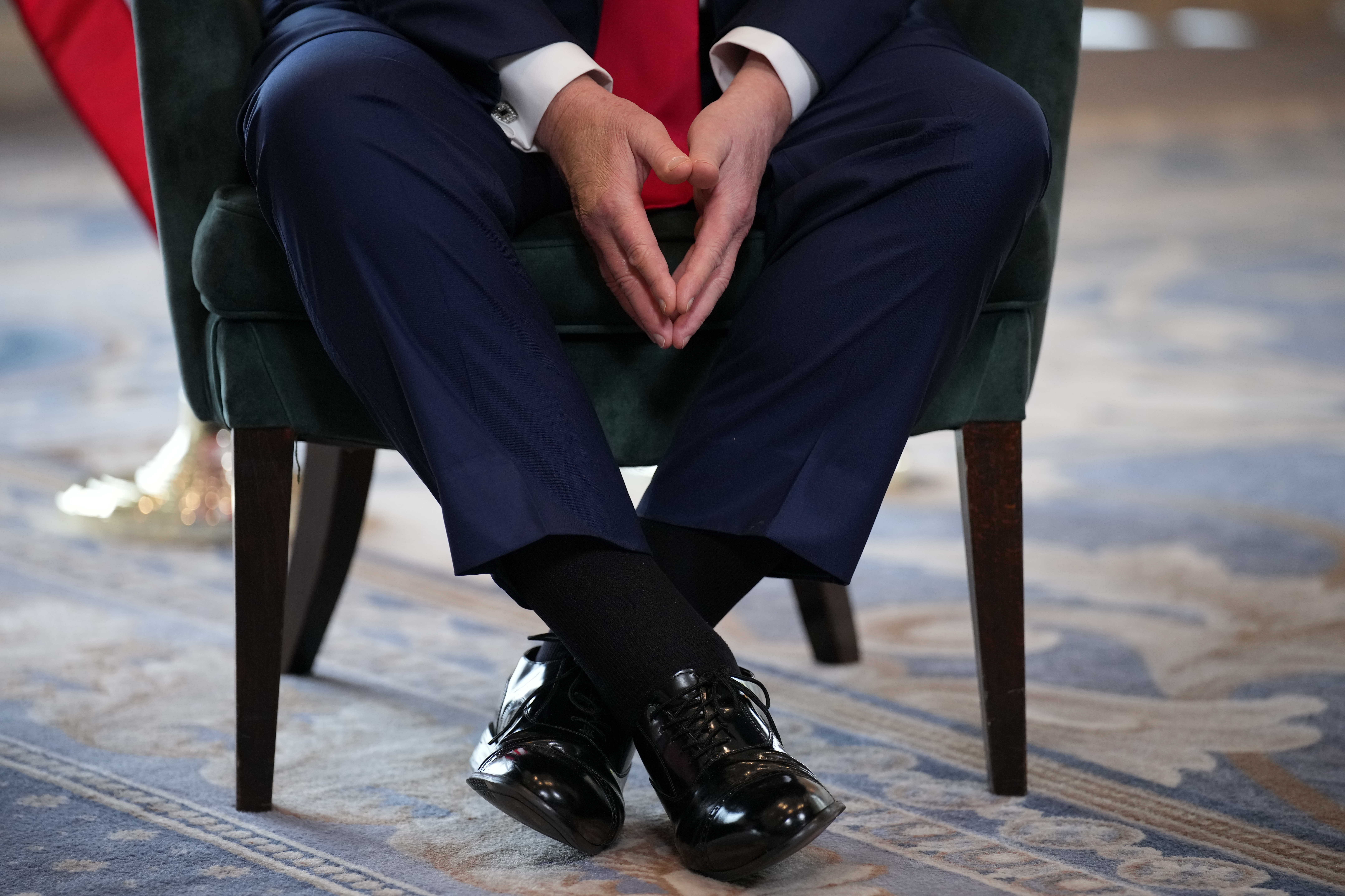 Donald Trump in a suit sits with hands clasped and legs crossed, showcasing polished shoes, in a formal setting, shown from the waste down