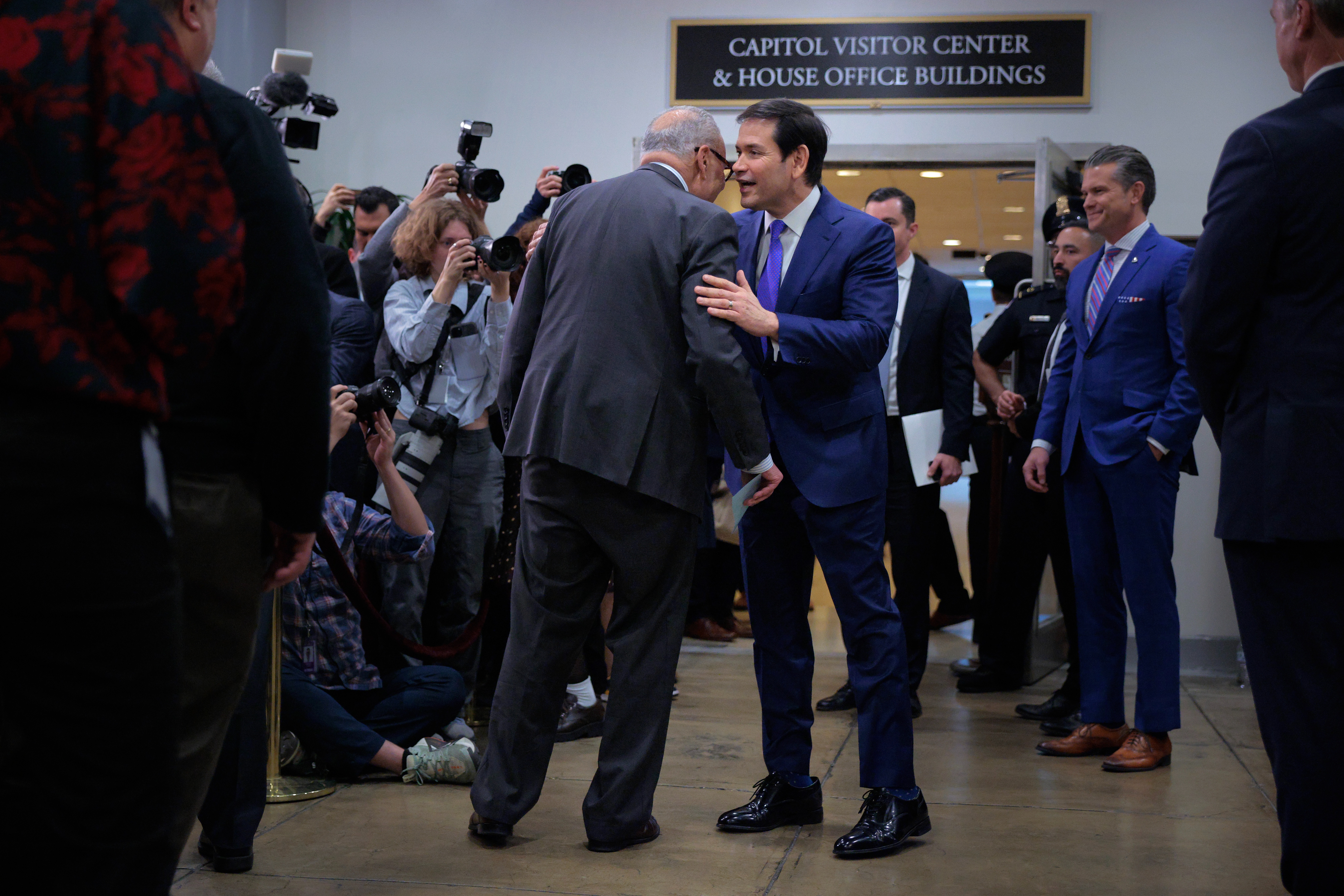 Marco Rubio leans in to speak with Chuck Schumer in a crowded hallway, with photographers capturing the moment