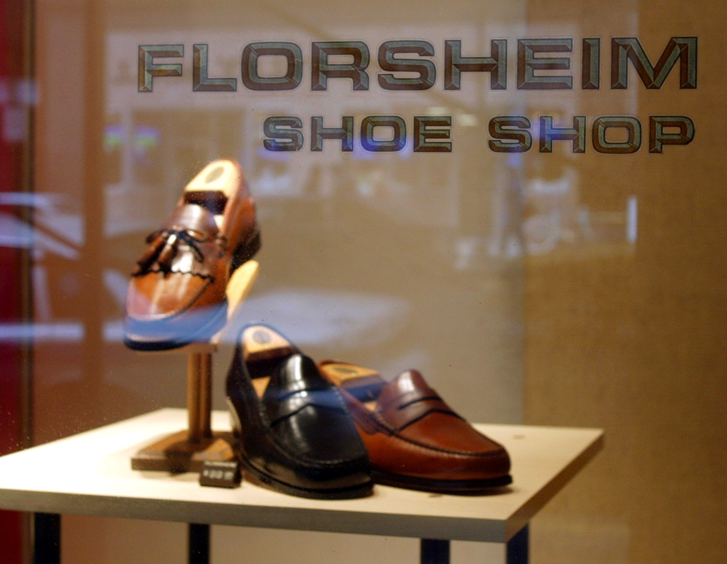 Storefront display featuring three pairs of men's leather shoes on a stand, viewed through a window with "Florsheim Shoe Shop" written on it