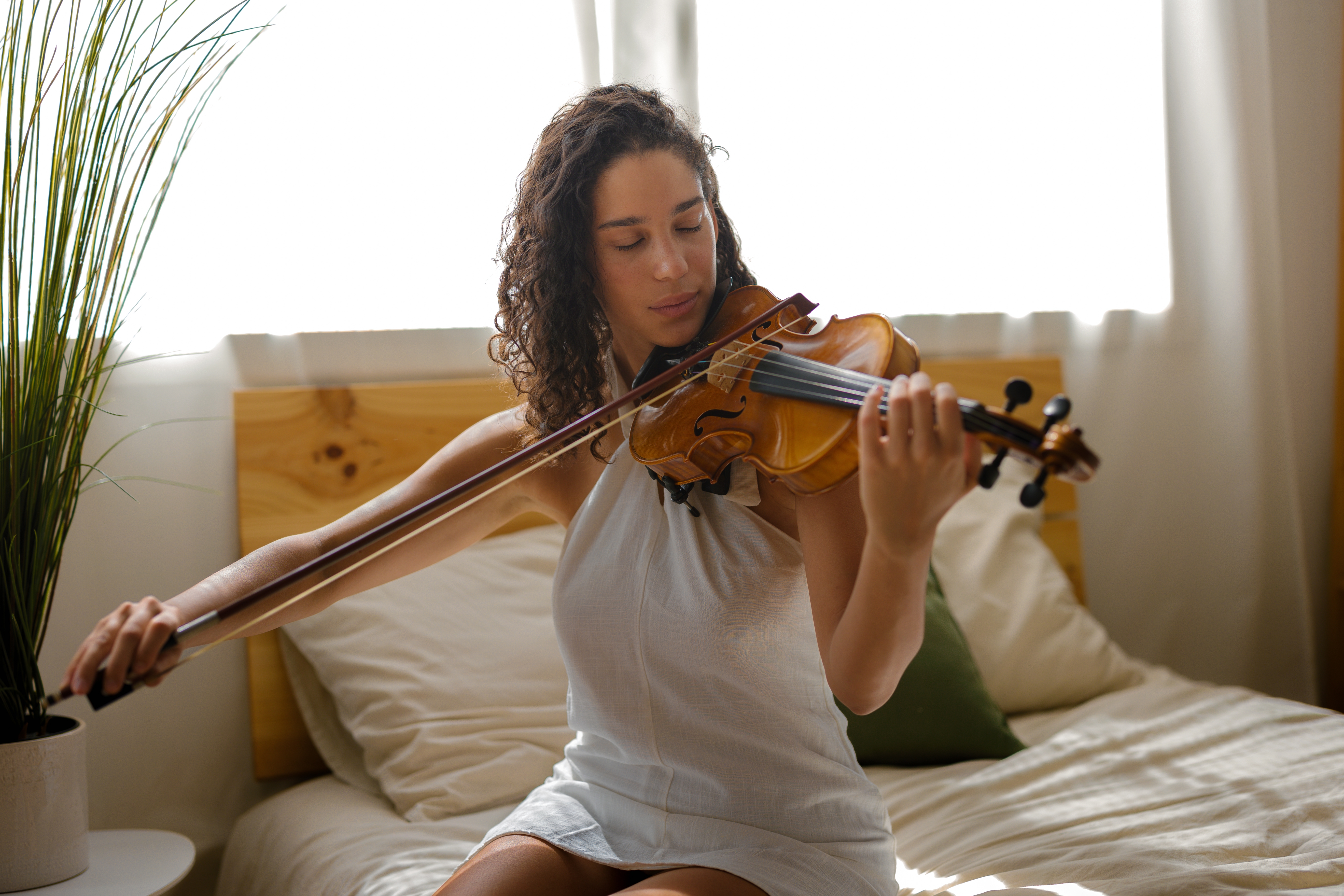 Woman plays violin connected  a bed, wearing a sleeveless dress, with sunlight streaming done  windows down  her