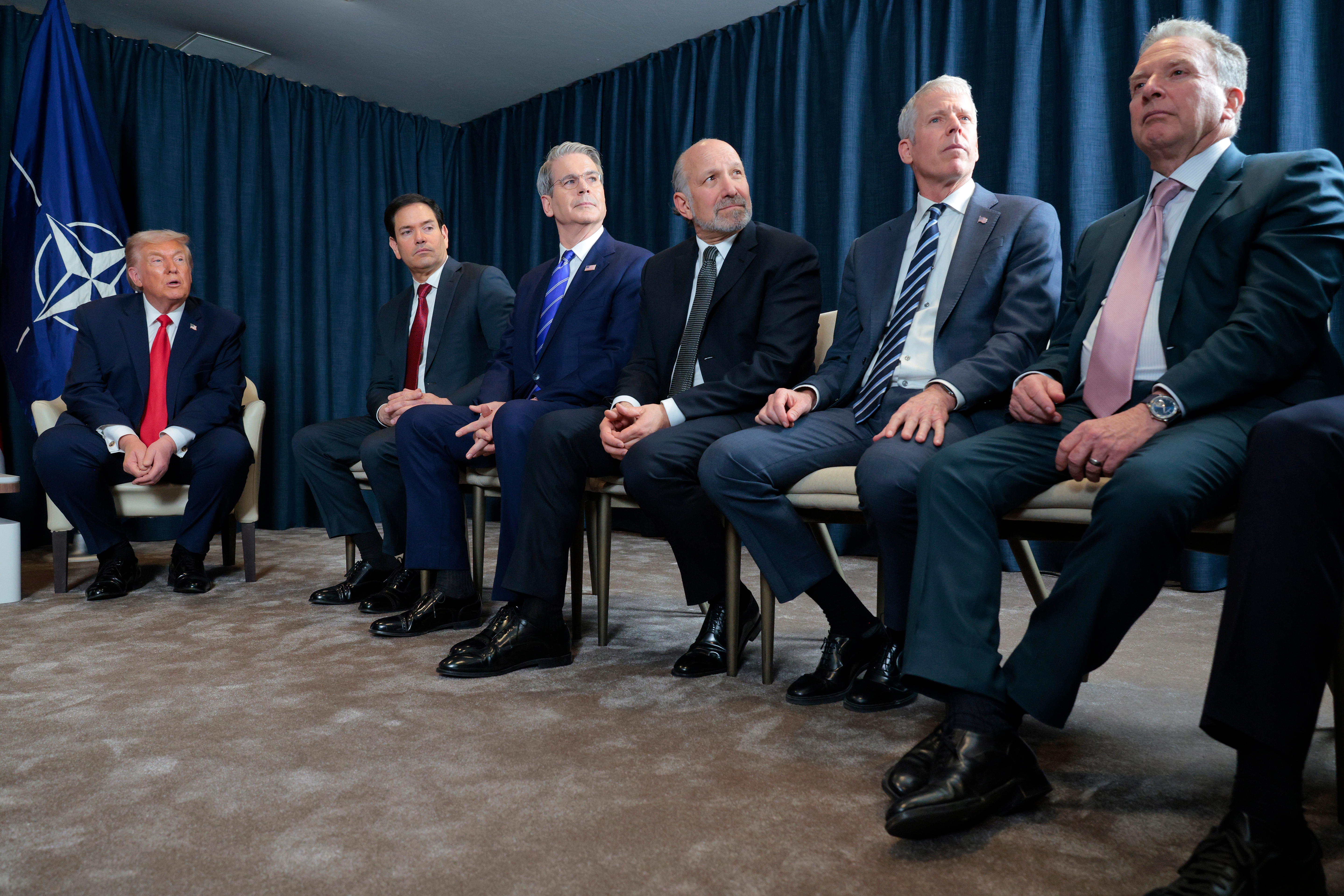 A group of men in suits sit on chairs in a formal setting with a NATO flag visible in the background