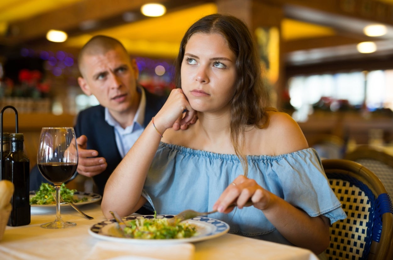 A woman looks uninterested as a man talks to her at a restaurant table. She gazes away, chin resting on her hand, while holding a fork