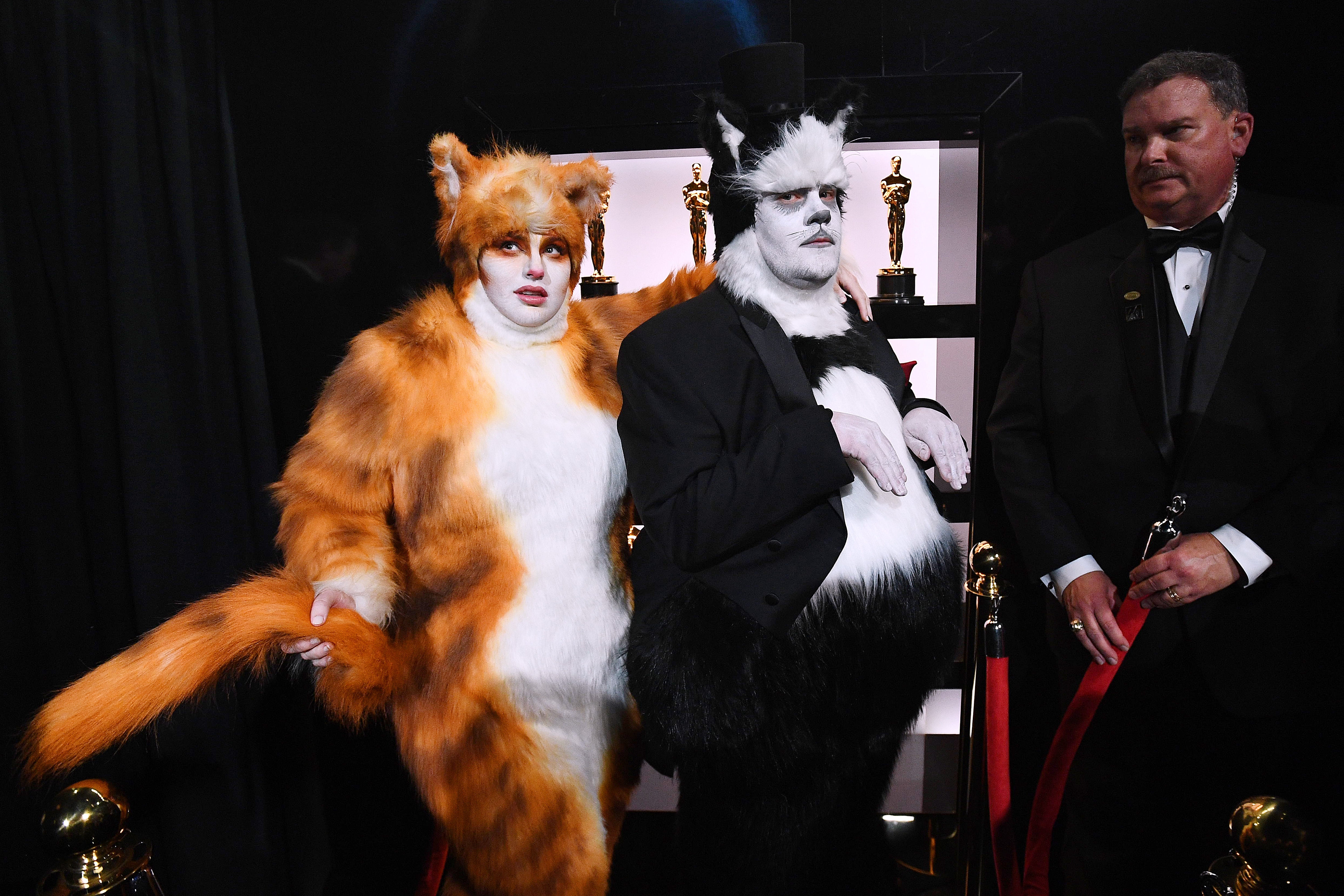 To personer i store myke kattekostymer poserer på en rød løper, som ligner karakterer fra en teaterforestilling eller film. En mann i smoking står i nærheten