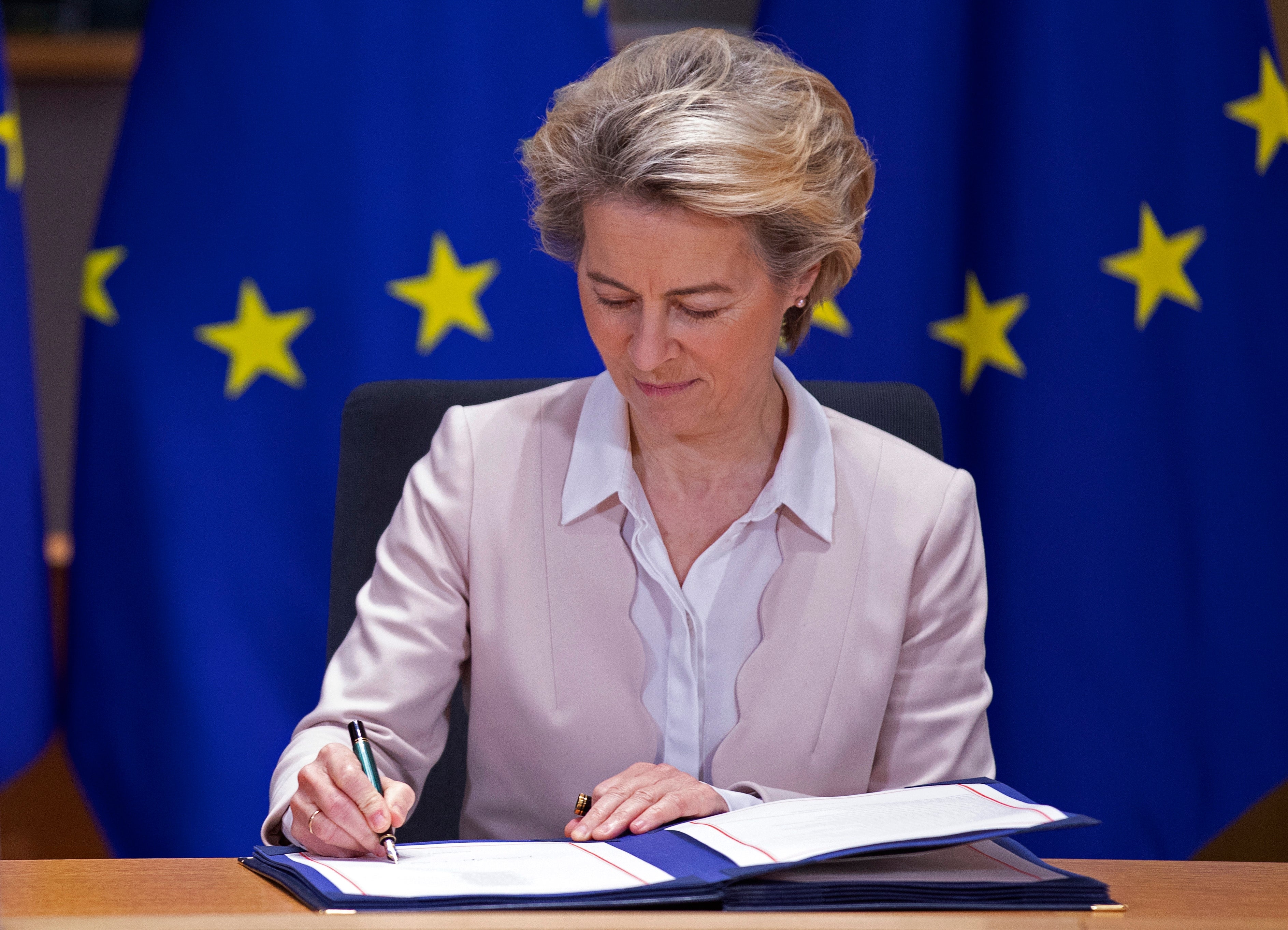 Person signerer et dokument ved et skrivebord med EU-flagg i bakgrunnen, iført en formell lys dress og hvit skjorte